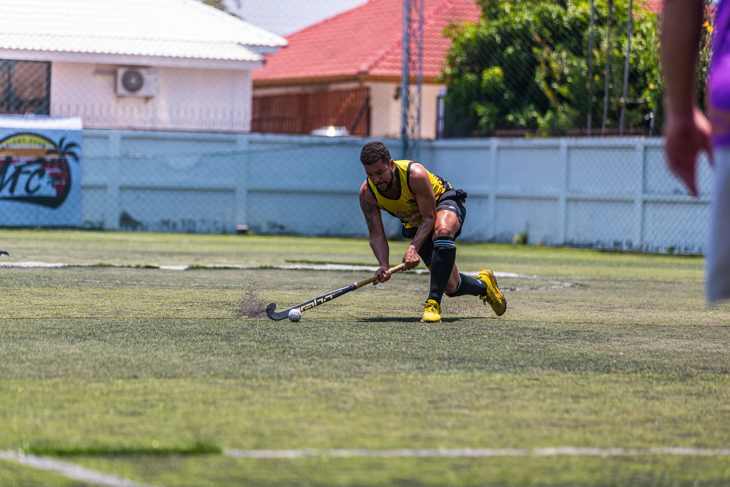 Romano Theunissen playing field hockey on a grassy field, wearing a yellow jersey, black shorts, and yellow shoes, with other players partially visible nearby, surrounded by a fence and buildings in the background.