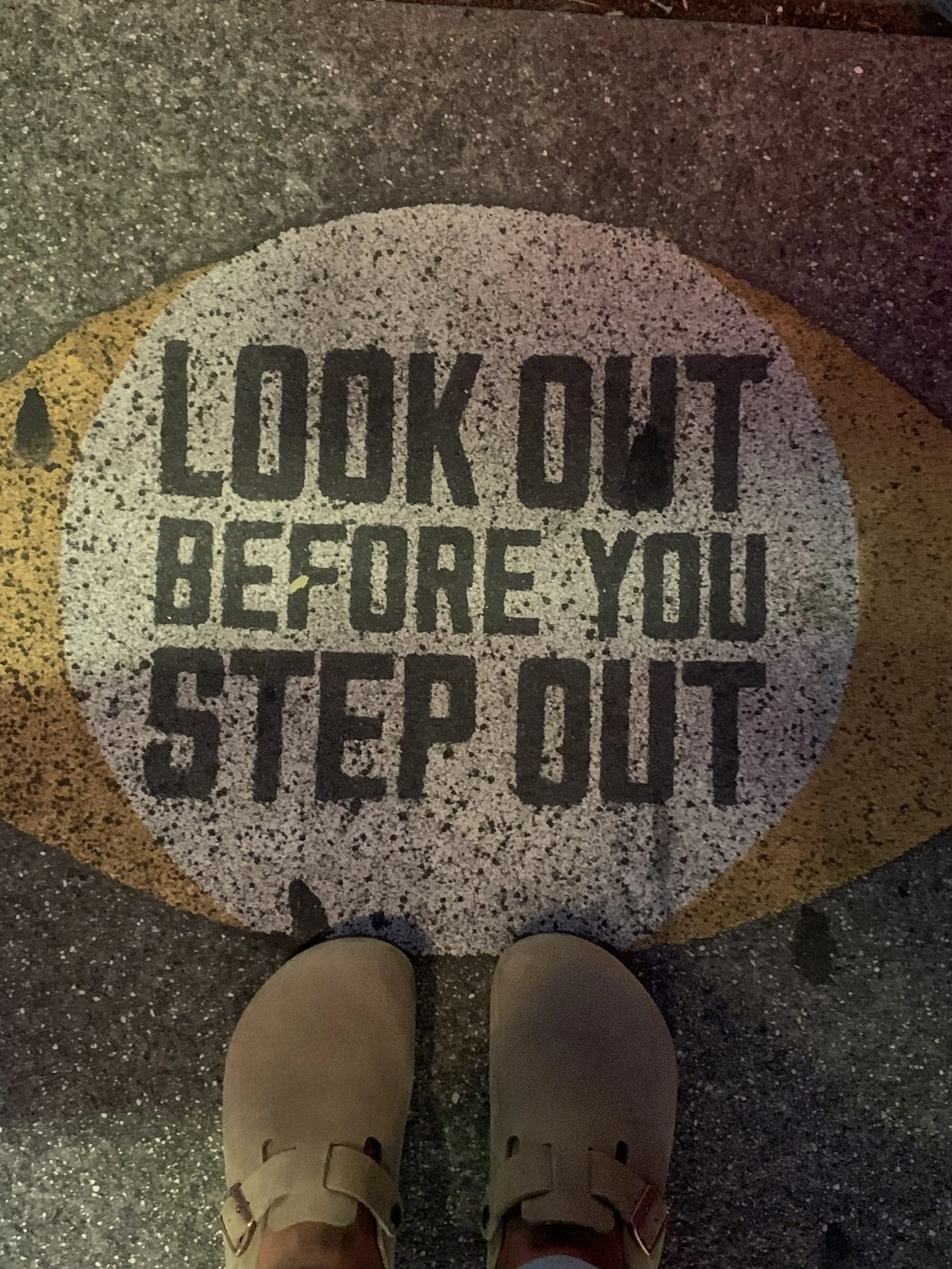 Footwear standing on a sidewalk with a circular sign that reads "LOOK OUT BEFORE YOU STEP OUT."