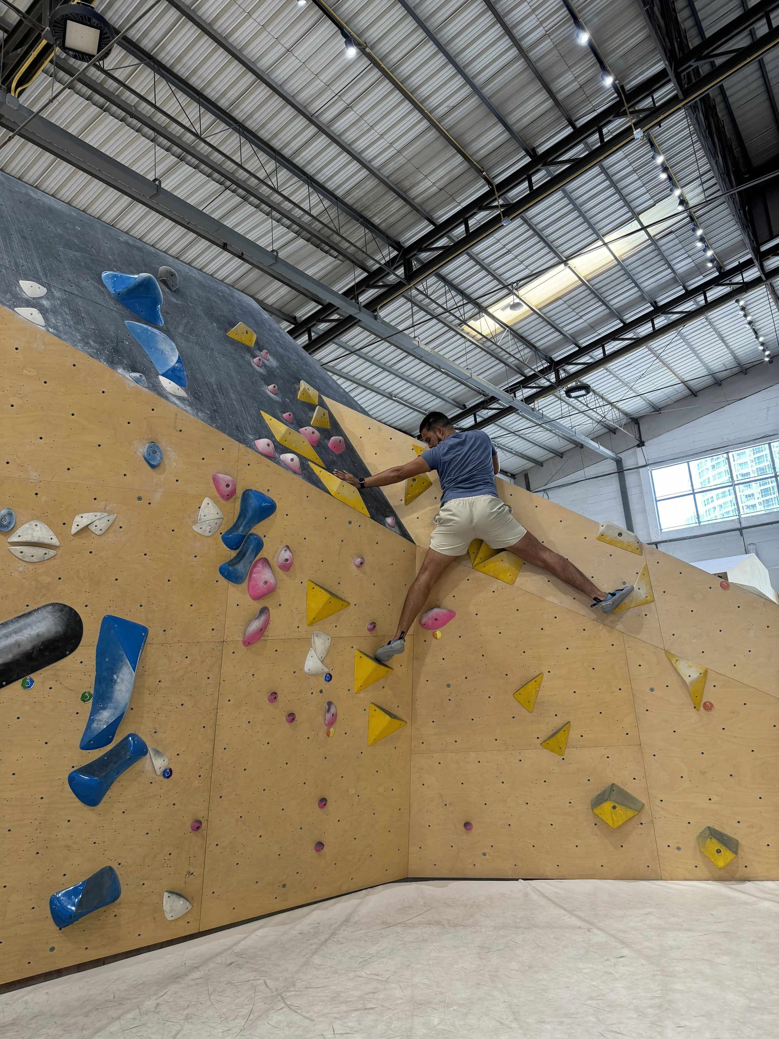 Romano Theunissen climbing an indoor bouldering wall with colorful holds, in a large gym with a metal ceiling and windows.