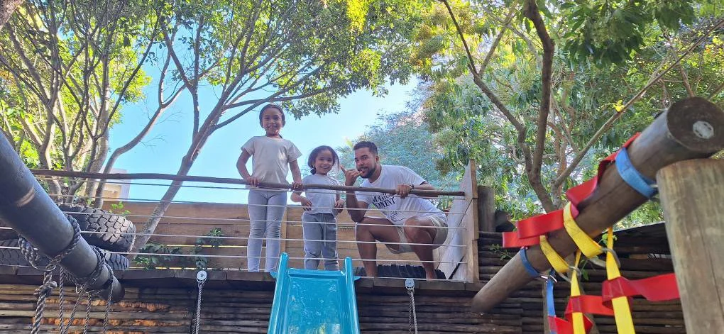 Romano Theunissen and his nieces at a playground, standing behind a wooden fence with trees in the background.