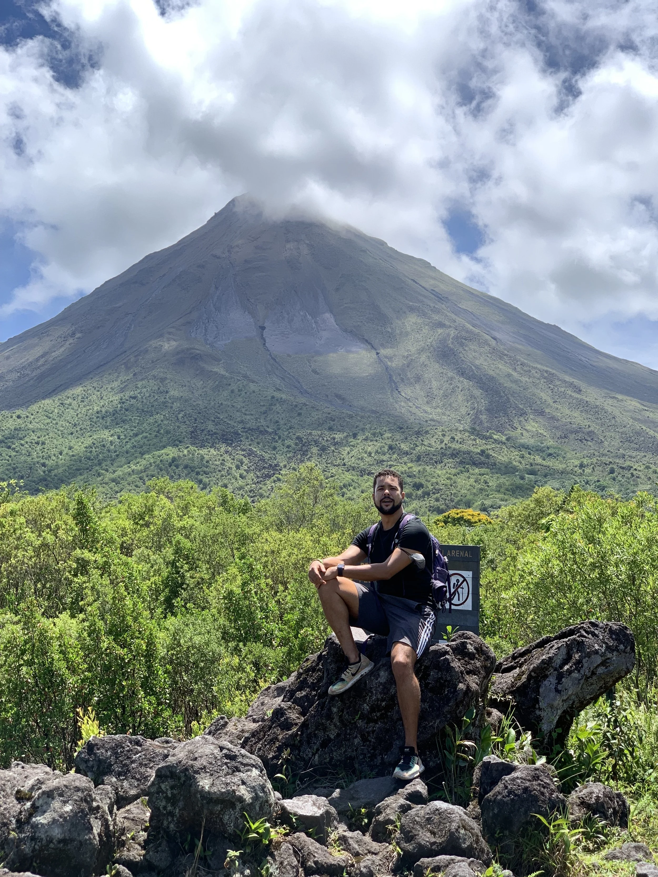 Romano Theunissen sitting on rocks in front of a lush green landscape with Arenal volcano in Costa Rica in the background, partially covered by clouds.