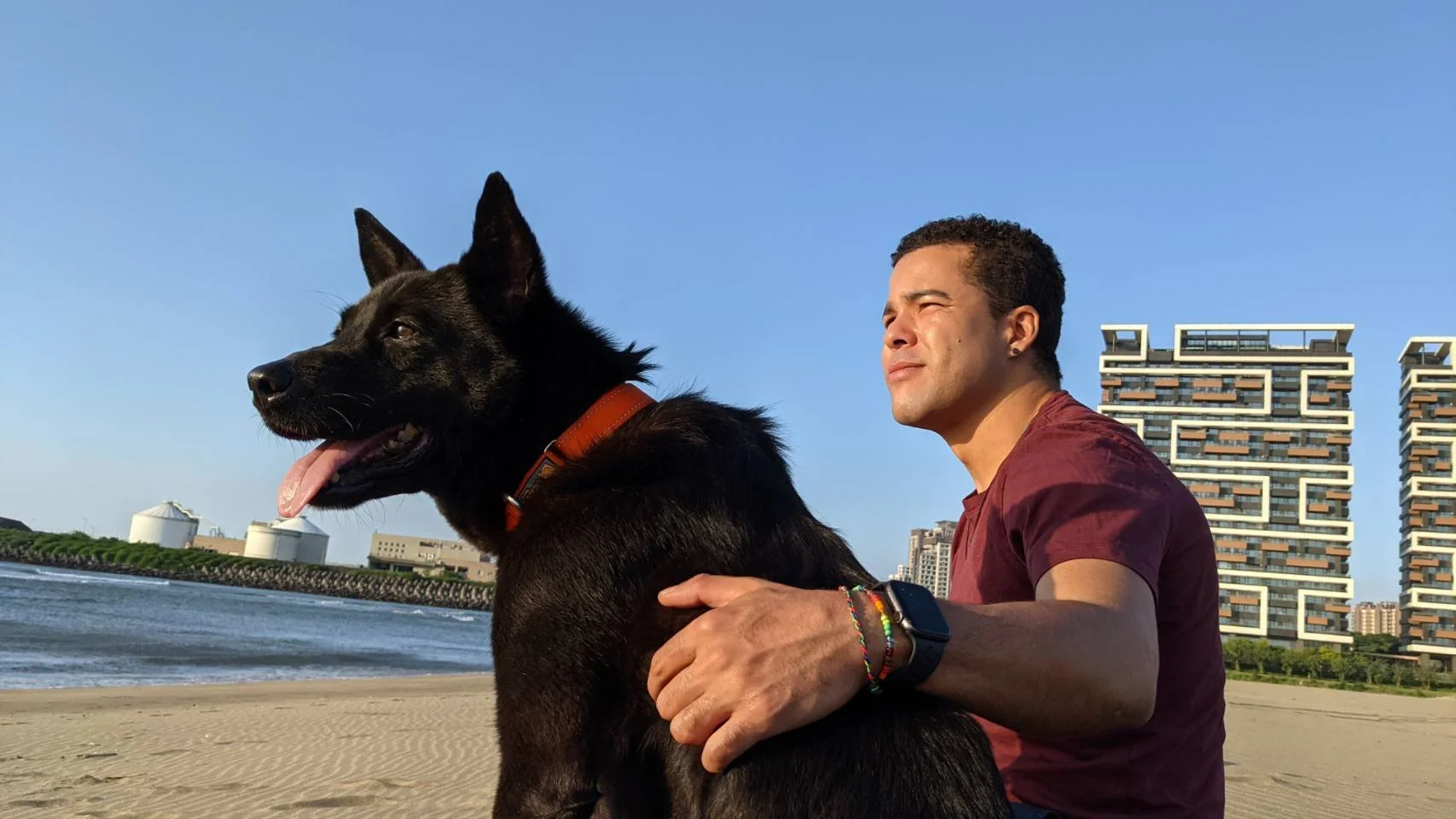 Romano Theunissen sitting on the beach with his black German shepherd dog, both facing towards the water with modern buildings in the background.