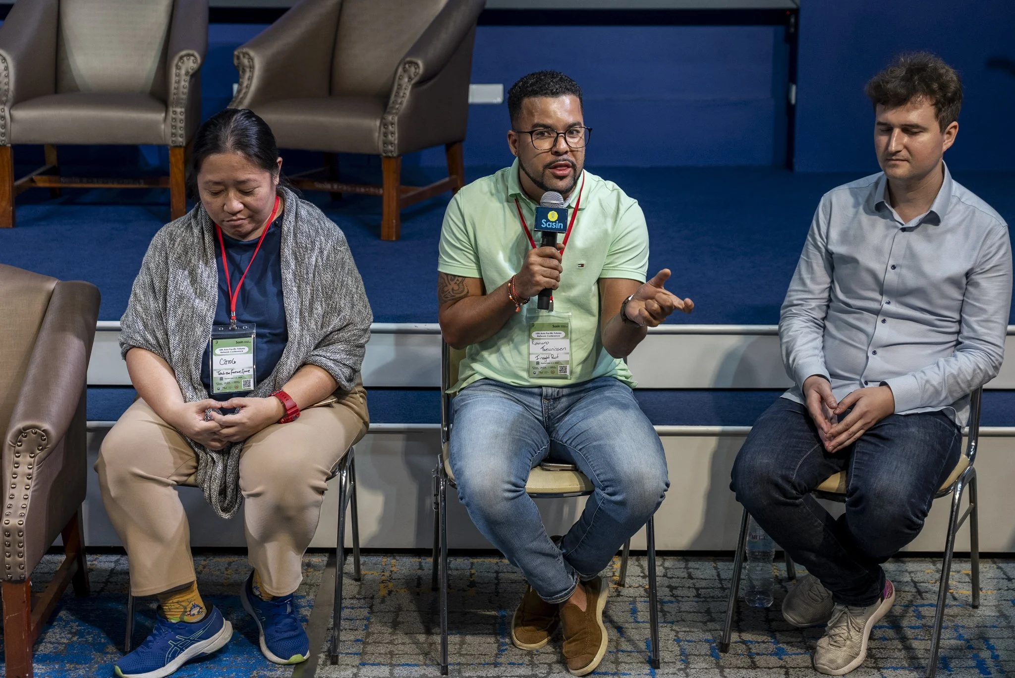 Three people sitting on chairs during a panel or discussion. Romano Theunissen is in the middle is speaking into a microphone.