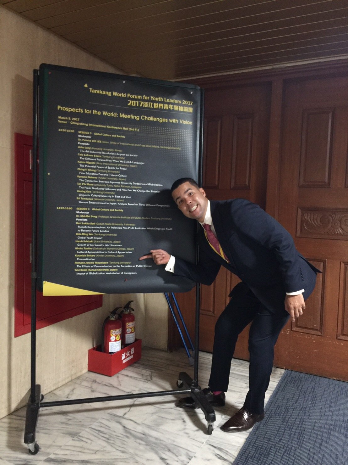 Romano Theunissen smiling and pointing at a conference schedule sign at the Tamkang World Forum for Youth Leaders 2017.