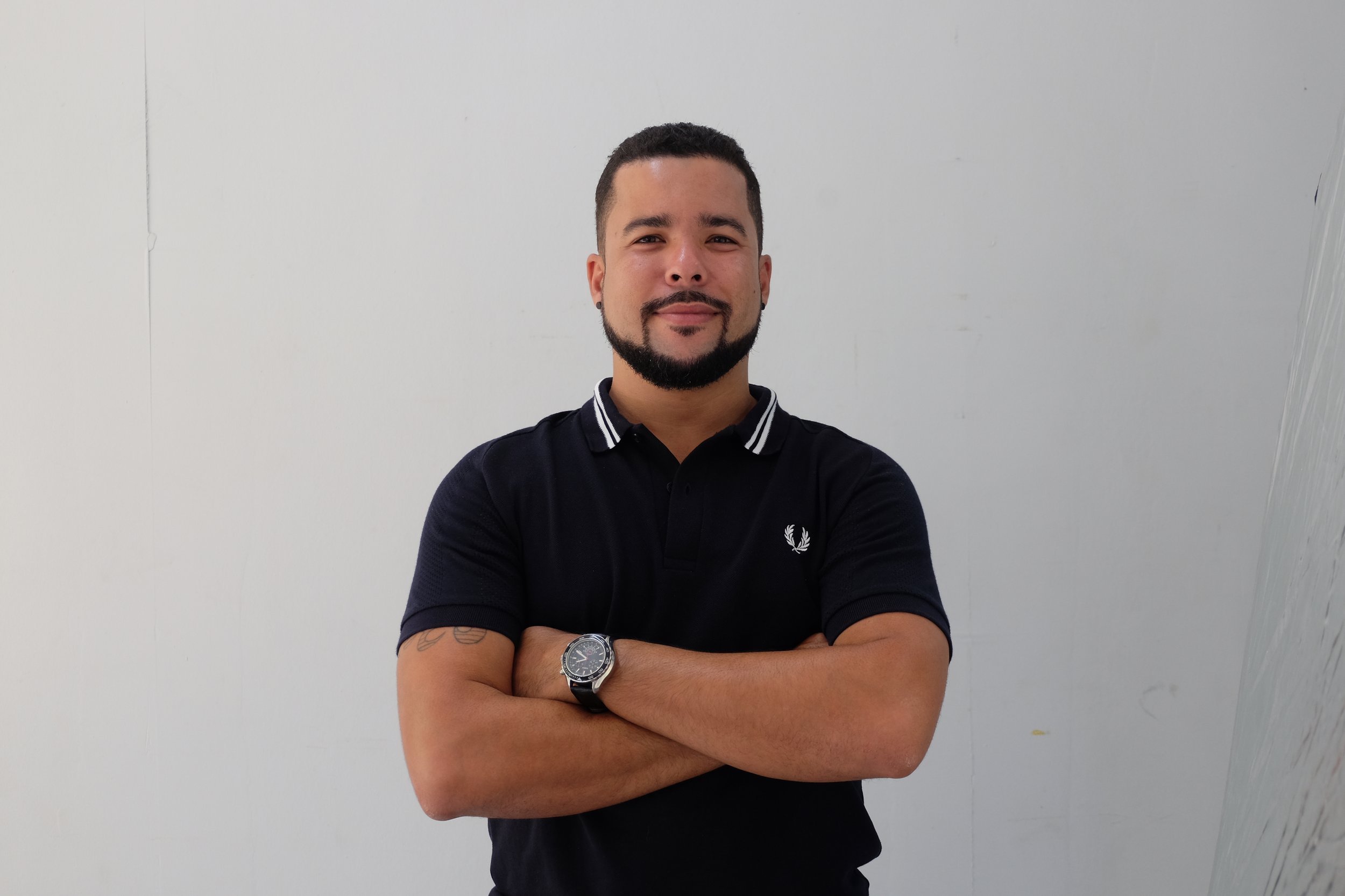This is Romano Theunissen's headshot. He is wearing a black polo shirt with white stripes on the collar, standing against a plain light-colored wall, with arms crossed and smiling.