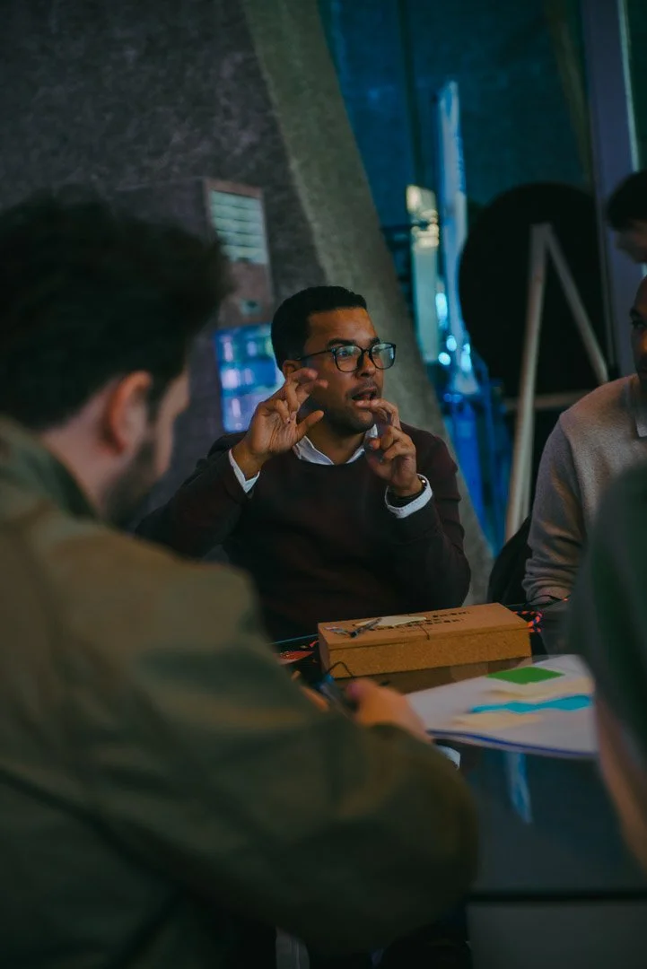 Romano Theunissen wearing glasses and a dark sweater is speaking at a table during a discussion. Other people are sitting around the table in a dimly lit room with a brick wall and some blue lighting.