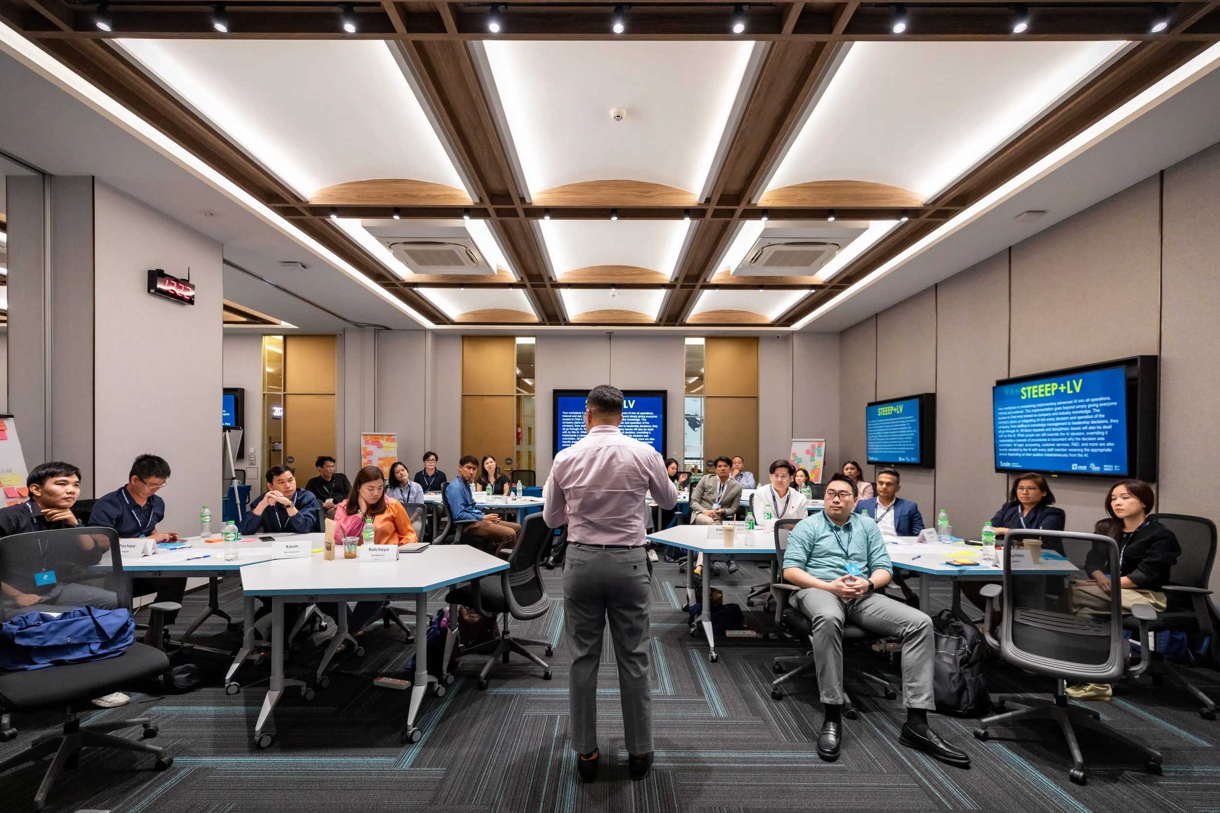 A diverse group of MBA students attending a lecture in a modern conference room, with Romano Theunissen standing in front of them and multiple screens displaying information.