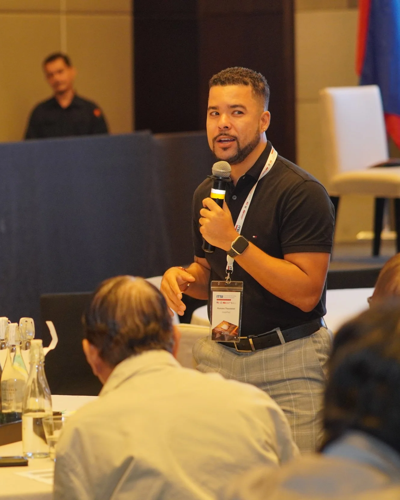 Romano Theunissen speaking into a microphone and facilitating at a workshop on semiconductors. He is wearing a black shirt, a watch, and a conference badge. In the background, there is another person standing and a secretarial desk or table.