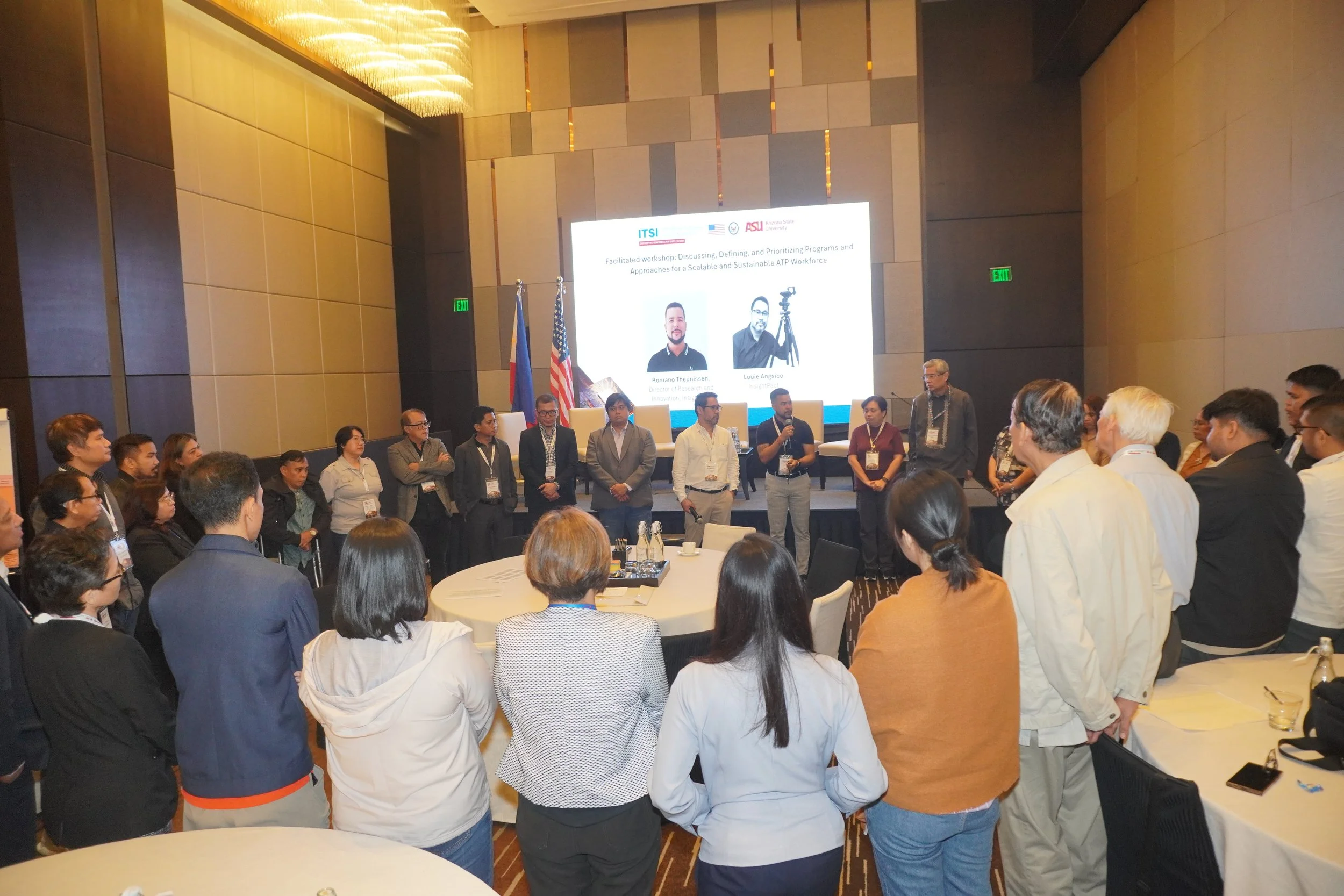Group of people standing in a circle in a conference room, listening to Romano Theunissen, with a large presentation screen displaying names, photos, and logos behind them.