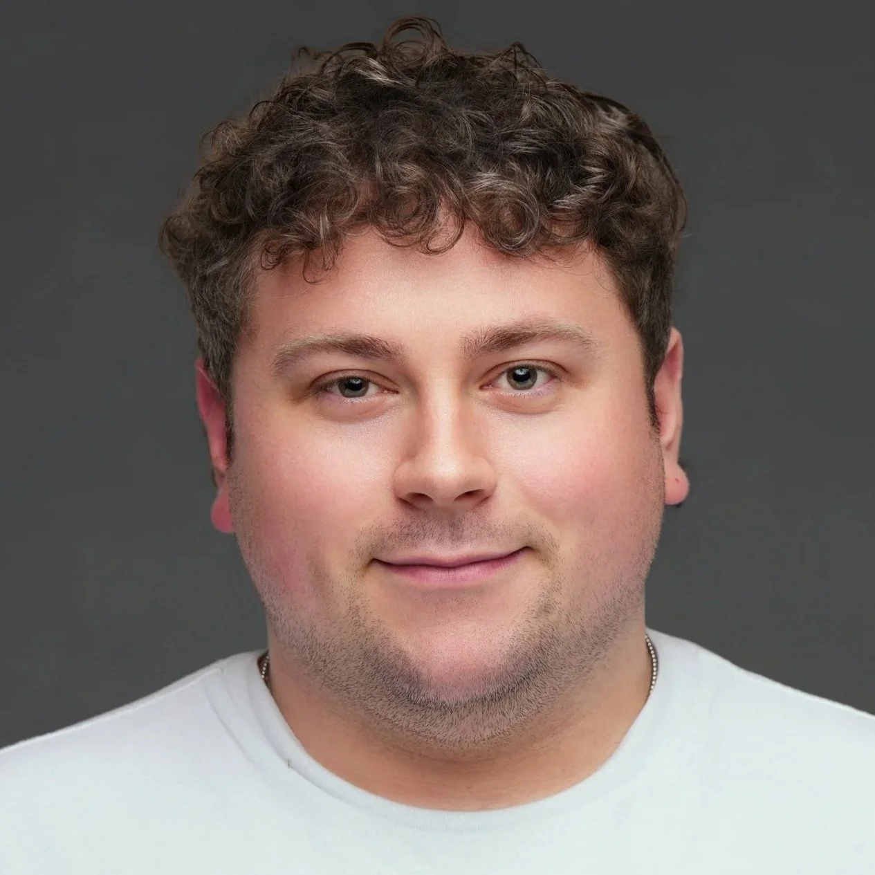 A young man with curly brown hair, blue eyes, and fair skin, smiling in a close-up portrait against a dark gray background.