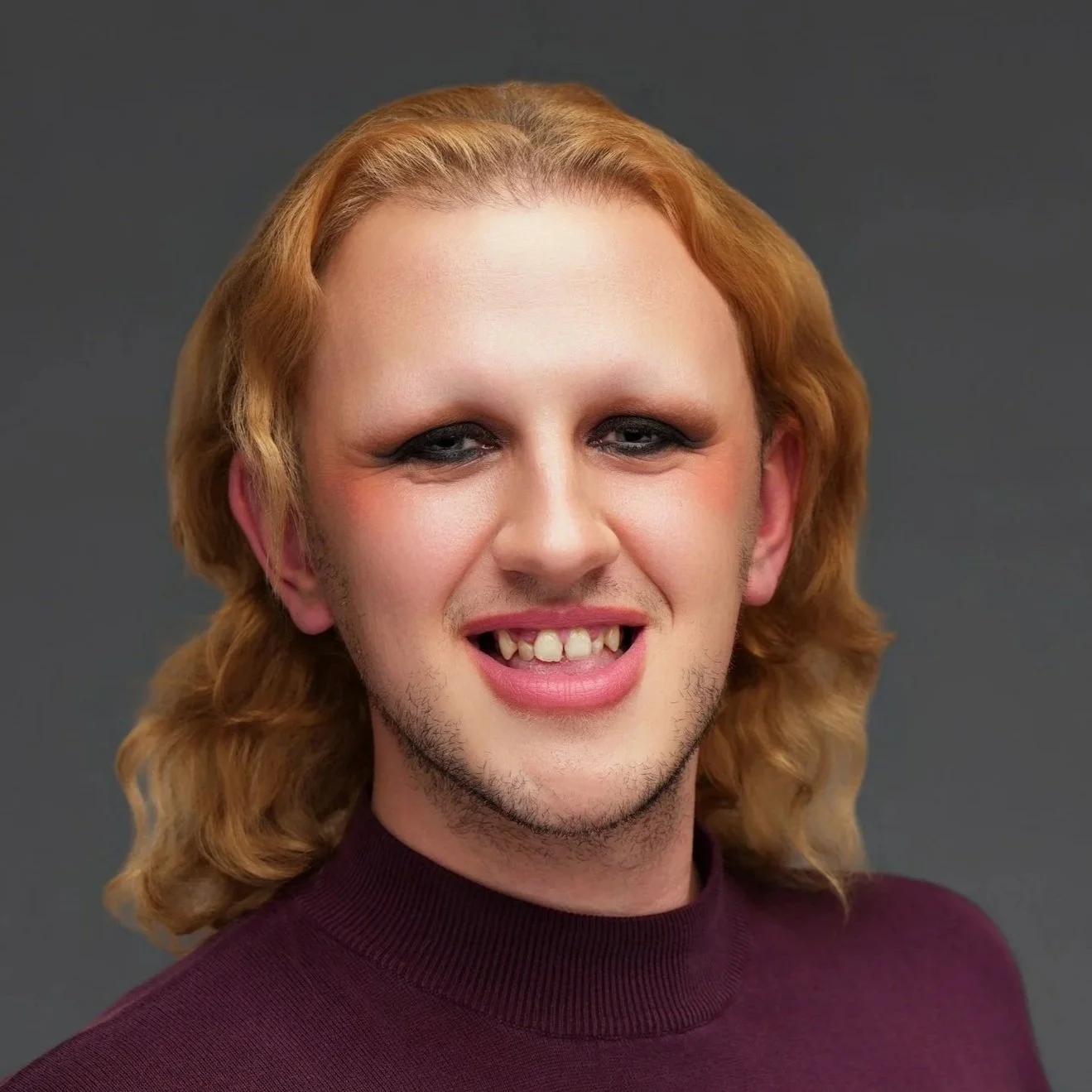 A person with long blonde hair, makeup, and a maroon turtleneck sweater, smiling with teeth showing, against a plain gray background.