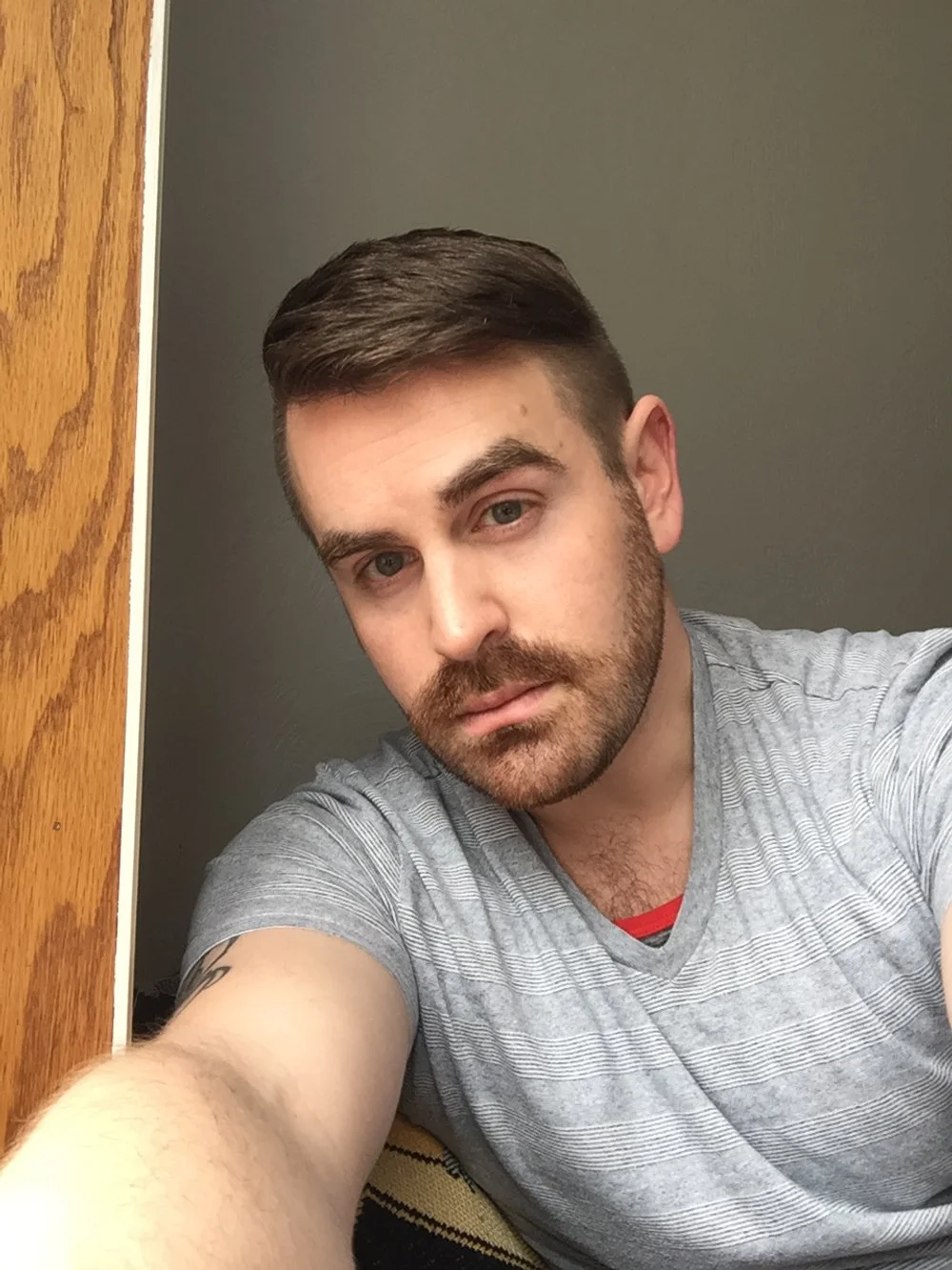 A young man with dark hair, beard, and mustache taking a selfie. He is wearing a gray striped t-shirt and is sitting in a room with a gray wall and a wooden door.