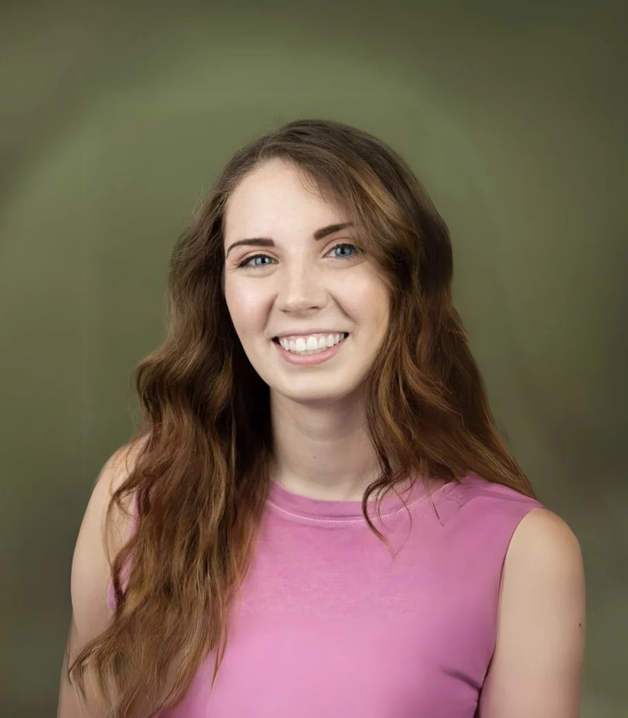 A young woman with long wavy brown hair, blue eyes, and fair skin, smiling at the camera, wearing a pink sleeveless top against a blurred green background.