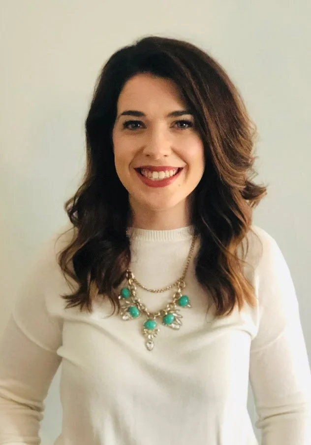 Woman with dark brown wavy hair smiling, wearing a white top and a statement necklace with turquoise beads.