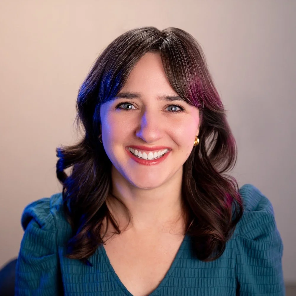 A smiling woman with wavy brown hair, wearing a teal top and gold earrings, in front of a neutral background.