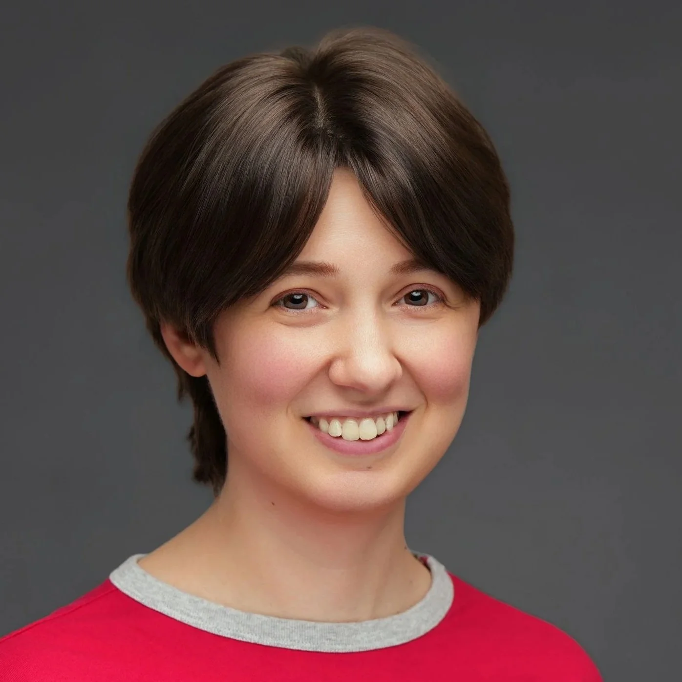 Portrait of a smiling person with short brown hair, wearing a red shirt with grey collar, against a grey background.
