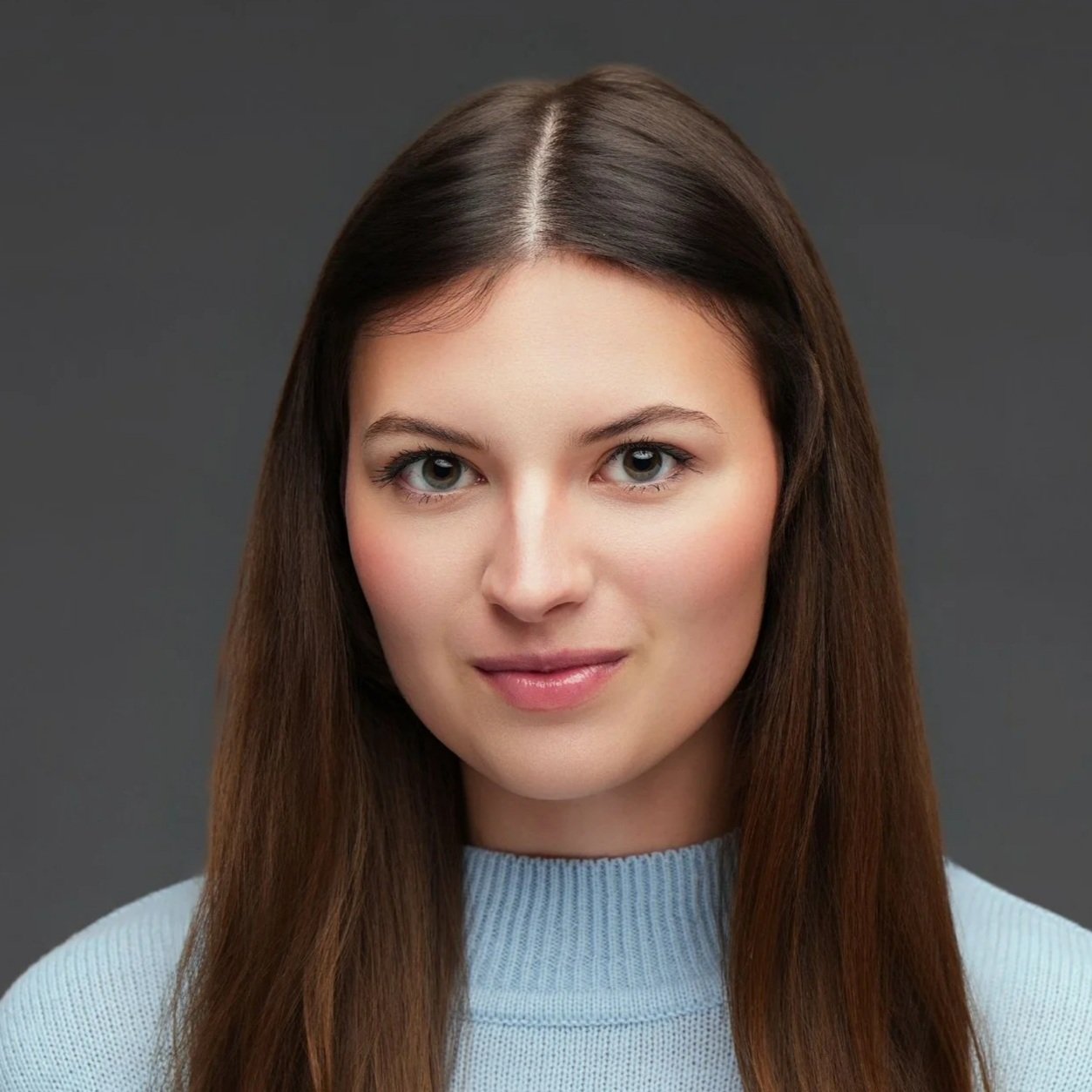 Headshot of a young woman with long brown hair, fair skin, and blue eyes, wearing a light blue sweater against a dark gray background.