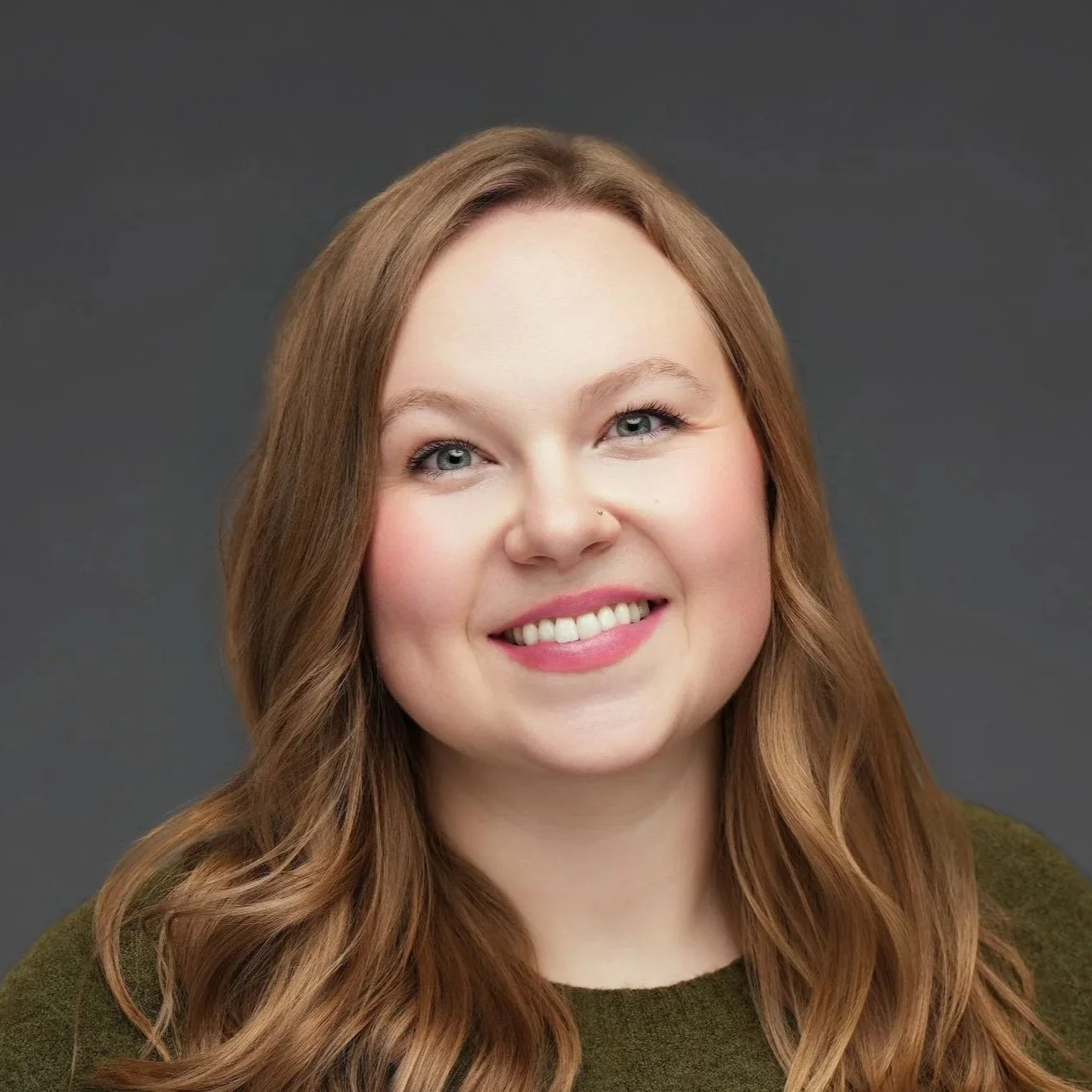 A young woman smiling with blonde hair, wearing a yellow top with blue and white accents, indoors with large windows in the background.