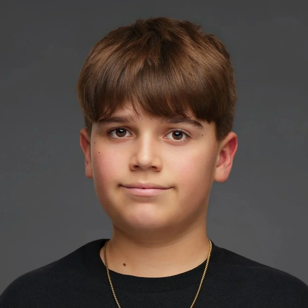Portrait of a young boy with brown hair, blue eyes, wearing a black shirt and a gold chain necklace, against a gray background.