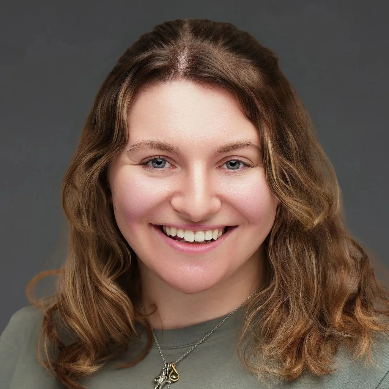 Close-up portrait of a young woman with shoulder-length wavy brown hair, blue eyes, and fair skin, smiling in front of a gray background.