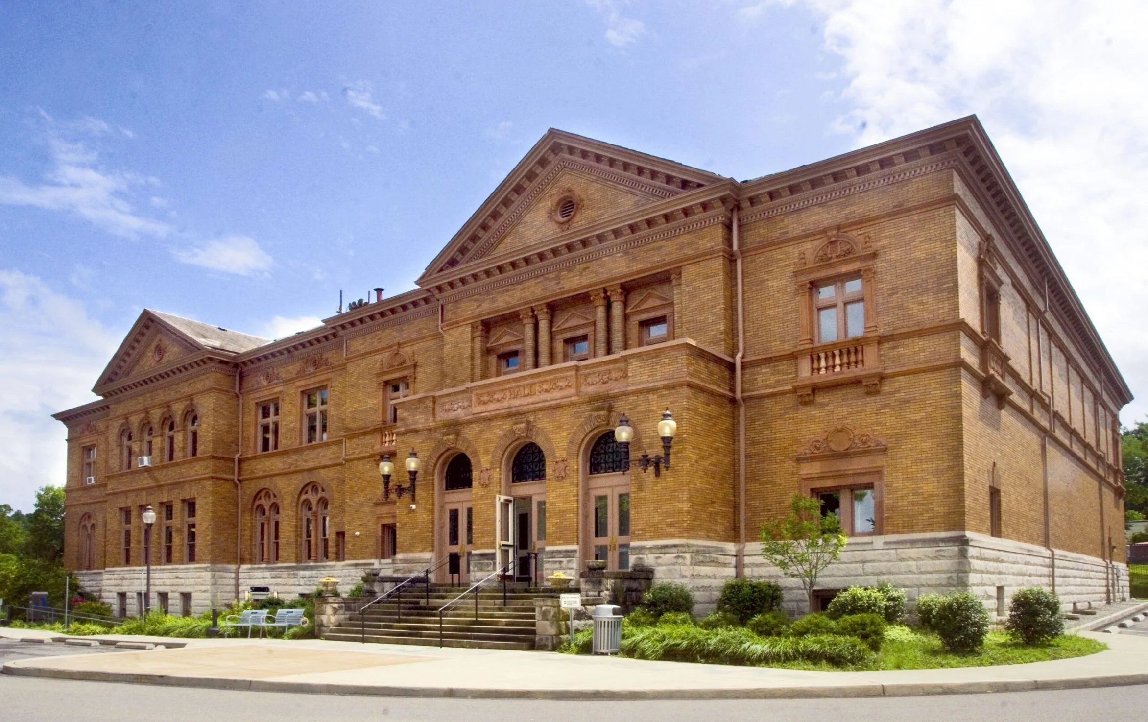 Historic brick building with steps leading up to entrance, surrounded by trees and street lamps, under a partly cloudy sky.