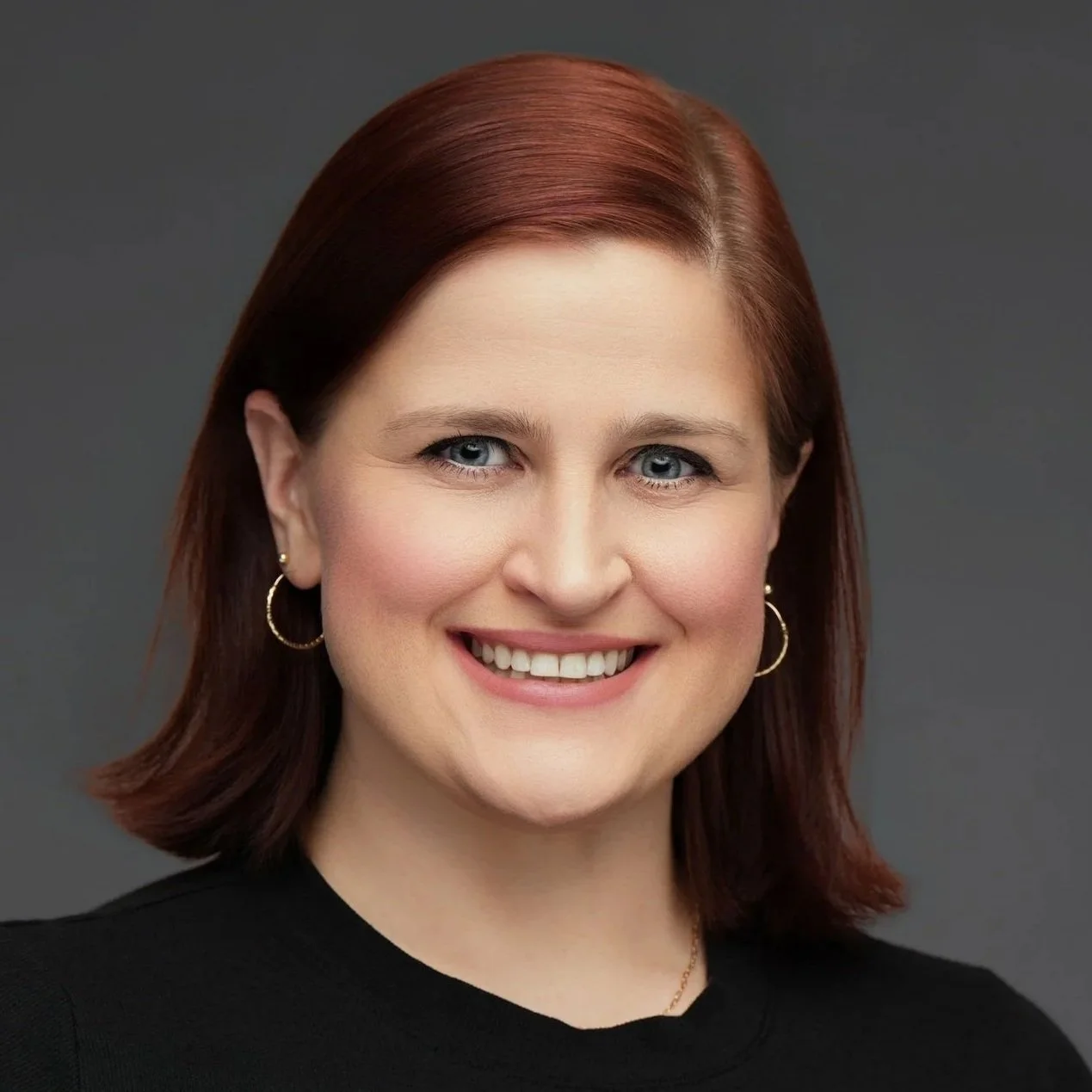 Close-up headshot of a woman with shoulder-length red hair, blue eyes, wearing hoop earrings and a black top, smiling against a gray background.