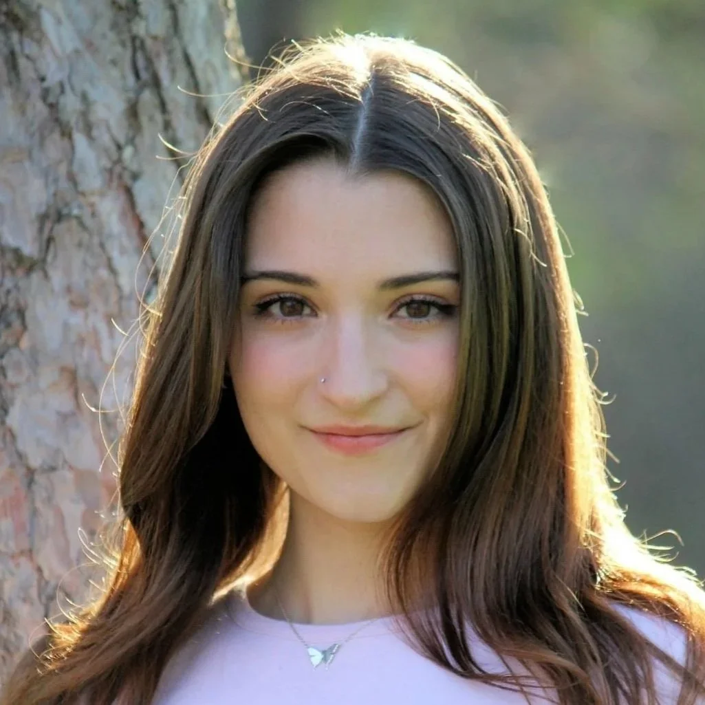 A young woman with long brown hair and a nose piercing smiling outdoors near a tree.