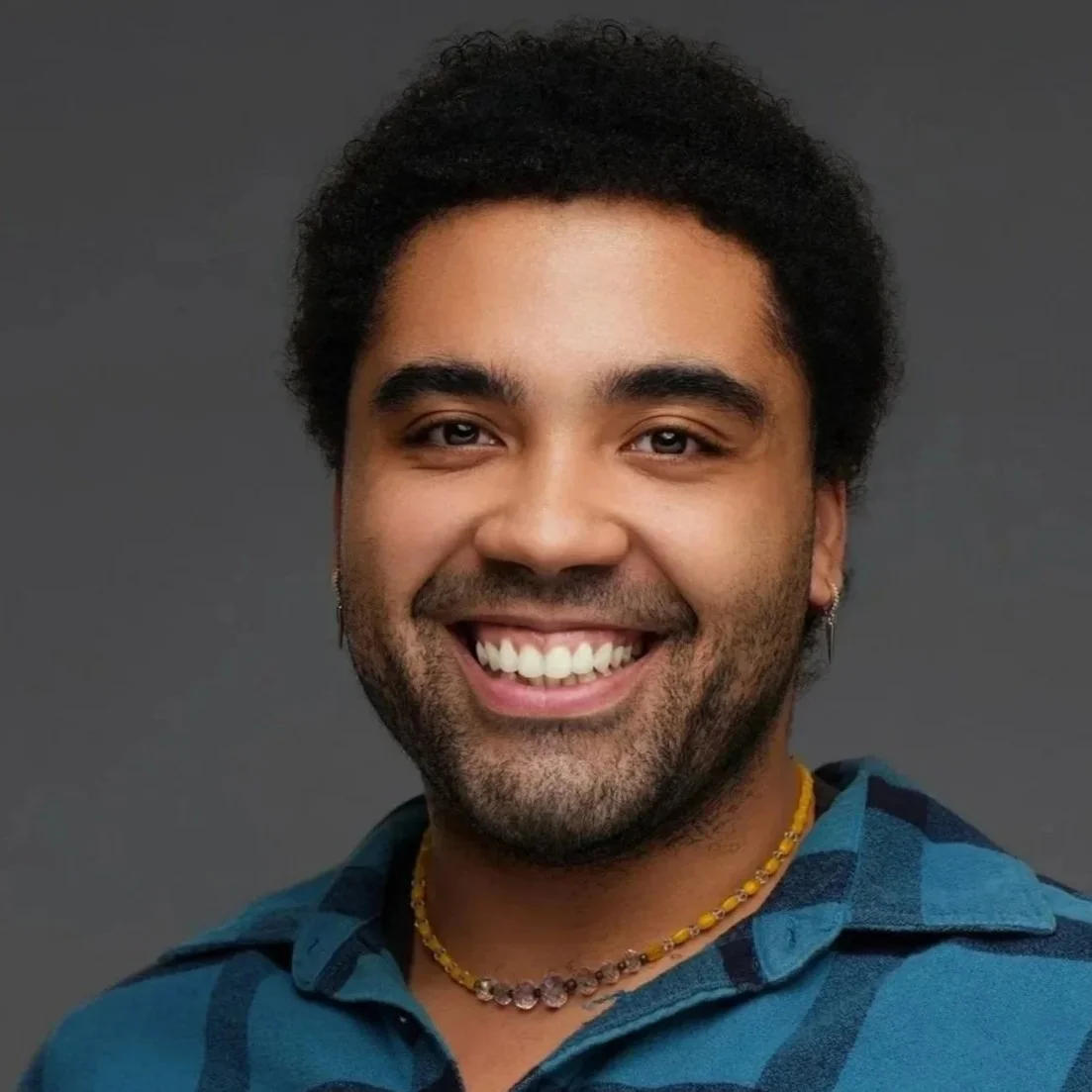 A smiling man with curly dark hair, wearing a blue checkered shirt, a yellow beaded necklace, and earrings, against a gray background.