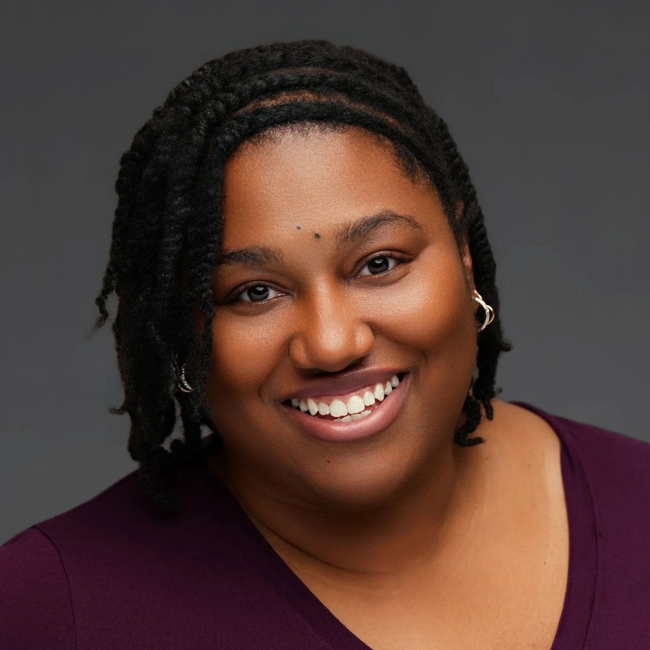 Close-up portrait of a smiling woman wearing her hair in twists, wearing a purple top and earrings, against a dark background.
