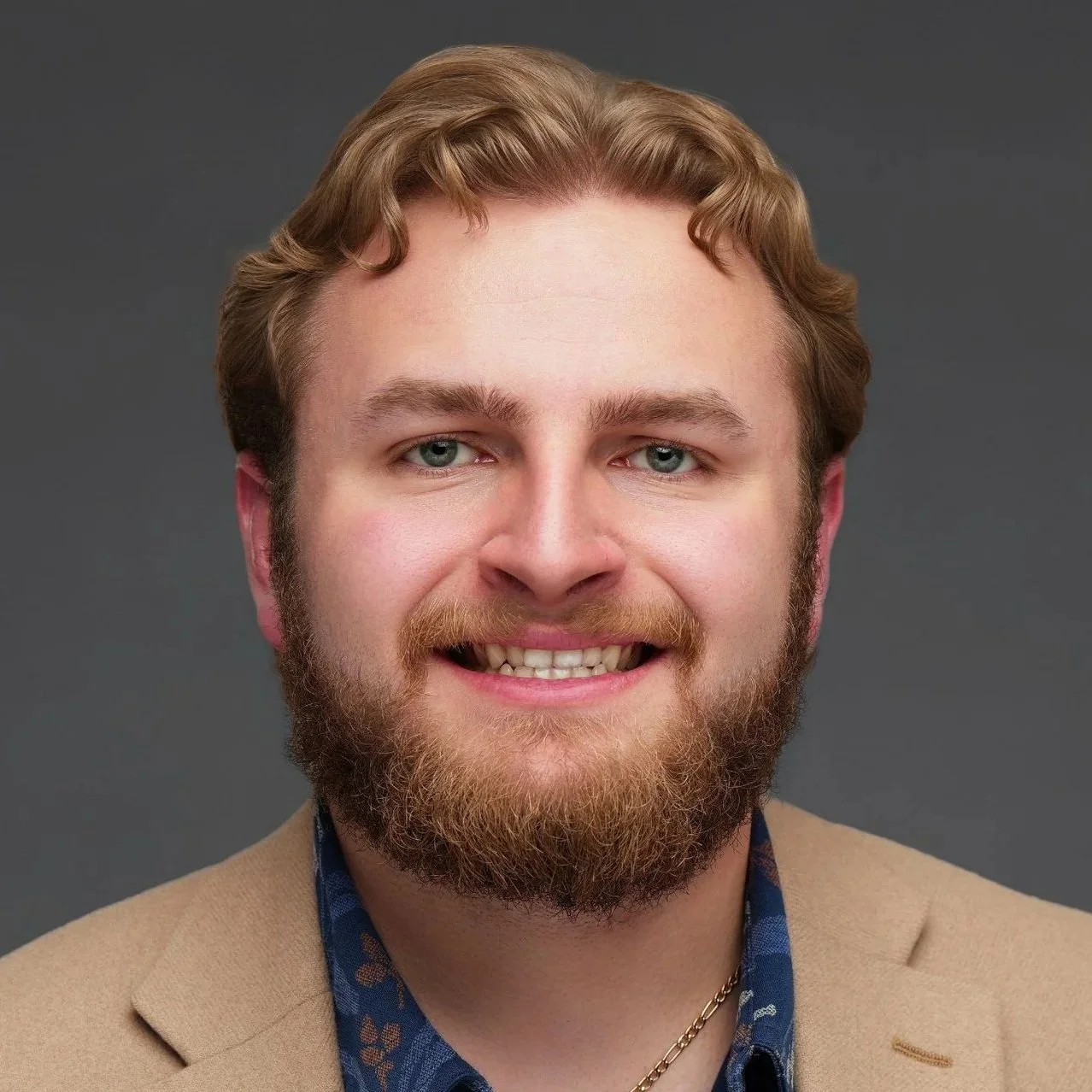 Headshot of a smiling man with a full beard, light brown hair, and blue eyes, wearing a beige blazer and blue patterned shirt against a dark gray background.