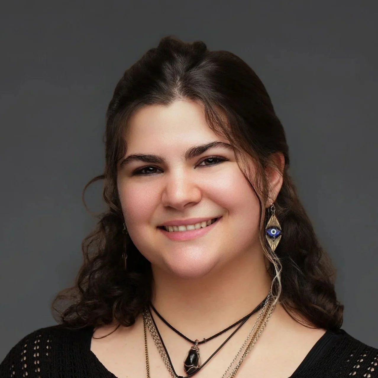 A young person with long curly brown hair, wearing earrings, necklaces, and a black top, smiling in front of a gray background.