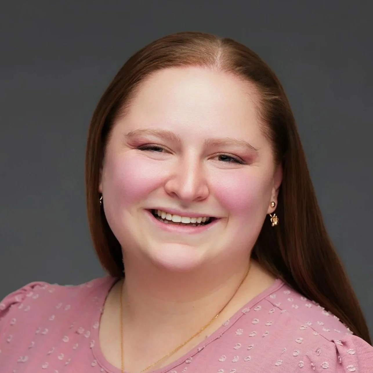 A smiling woman wearing a pink top, gold earrings, and a gold necklace against a gray background.