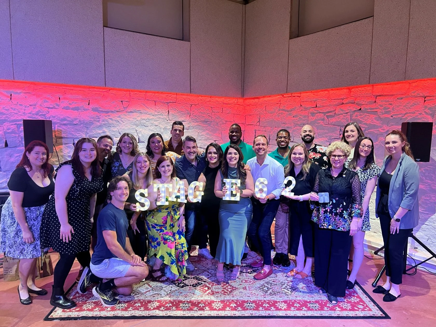 Group of people smiling in a celebratory setting holding letters and numbers spelling out 'Stage 62', with two large speakers in the background, on a patterned rug with a colorful stone wall backdrop and pink lighting.