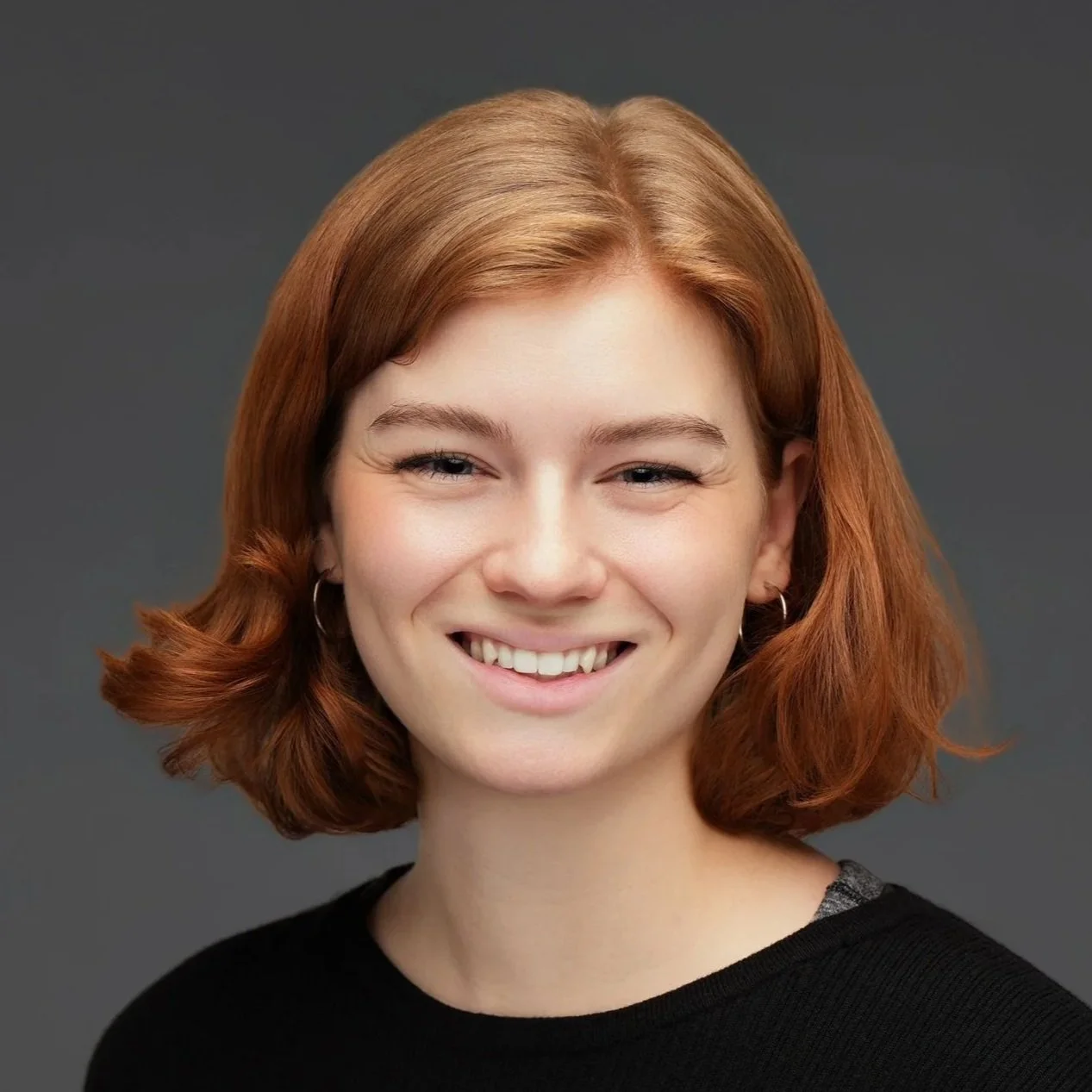 Portrait of a young woman with short reddish hair smiling against a dark gray background.
