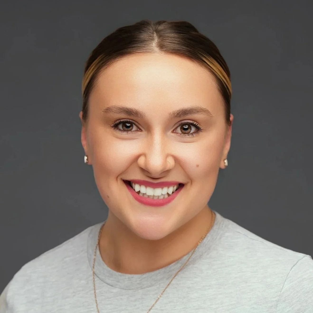 Close-up of a smiling woman with light brown hair styled back, wearing a gray top and small earrings, against a dark gray background.