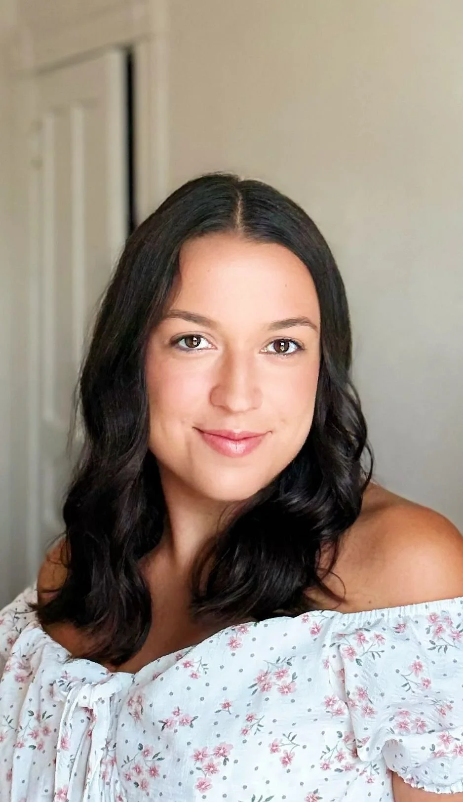 A young woman with shoulder-length dark hair, wearing a white off-the-shoulder floral top, smiling at the camera against a neutral background.