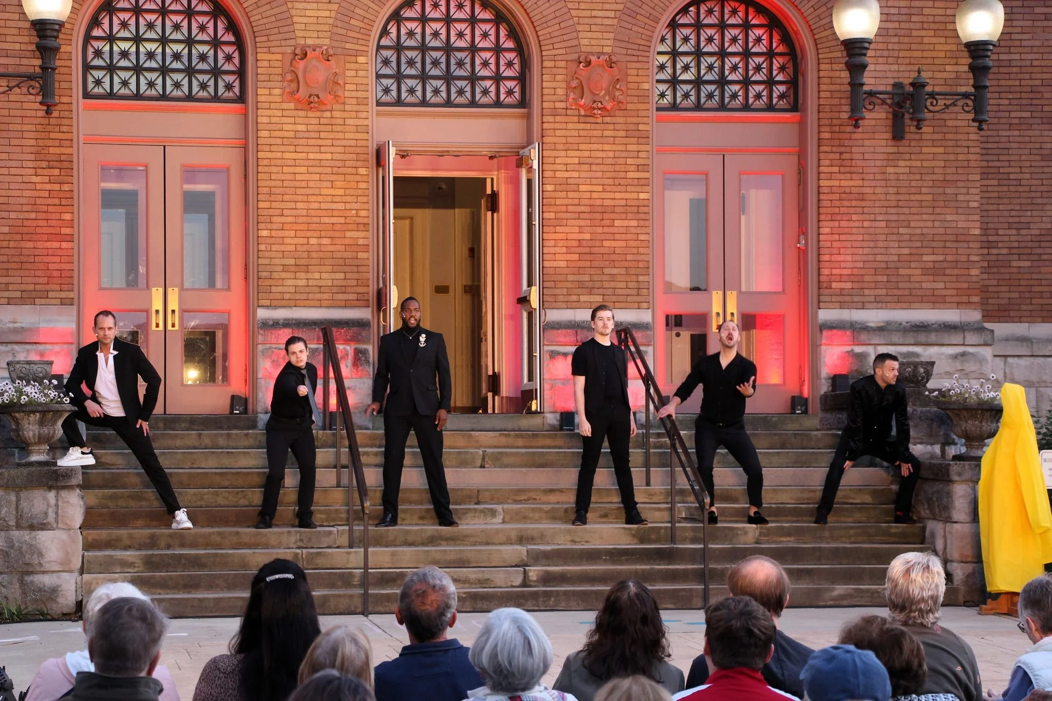 A group of six performers on steps in front of a brick building, engaging in a performance for an audience seated in front. The performers are dressed mostly in black, with some striking intense poses and expressions.