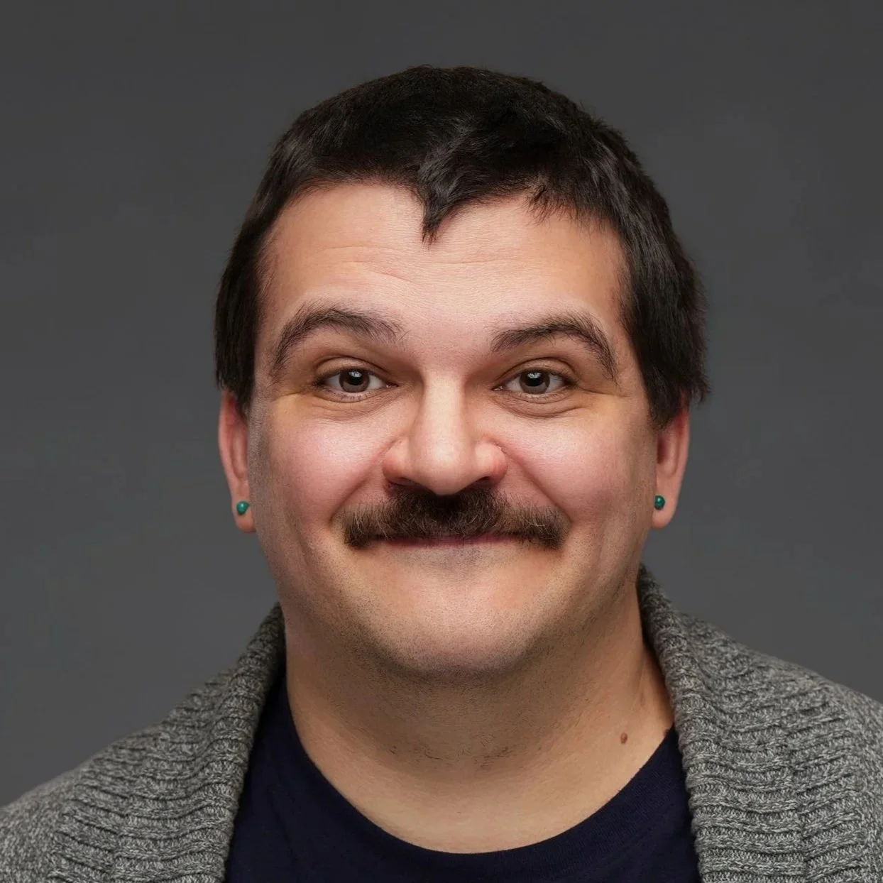 Close-up portrait of a man with short dark hair, mustache, light skin, wearing teal earrings, smiling, against a gray background.