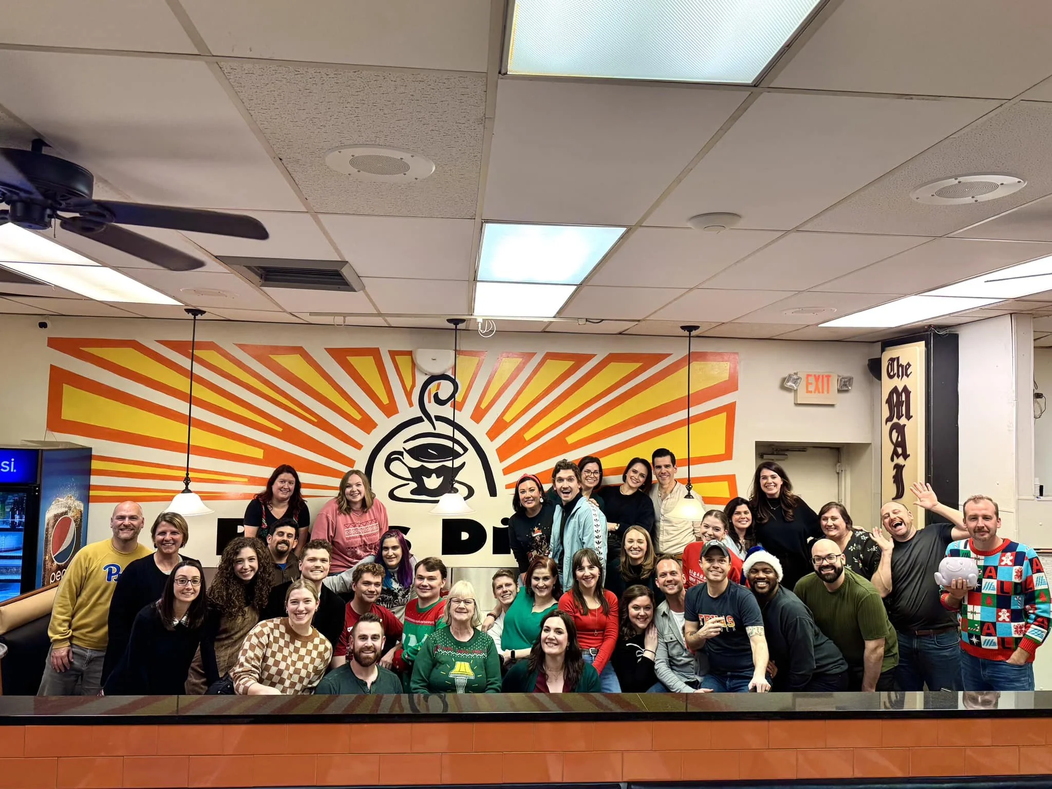 Group of people gathered together in front of a decorated wall with a coffee cup logo, celebrating indoors with some wearing festive attire.