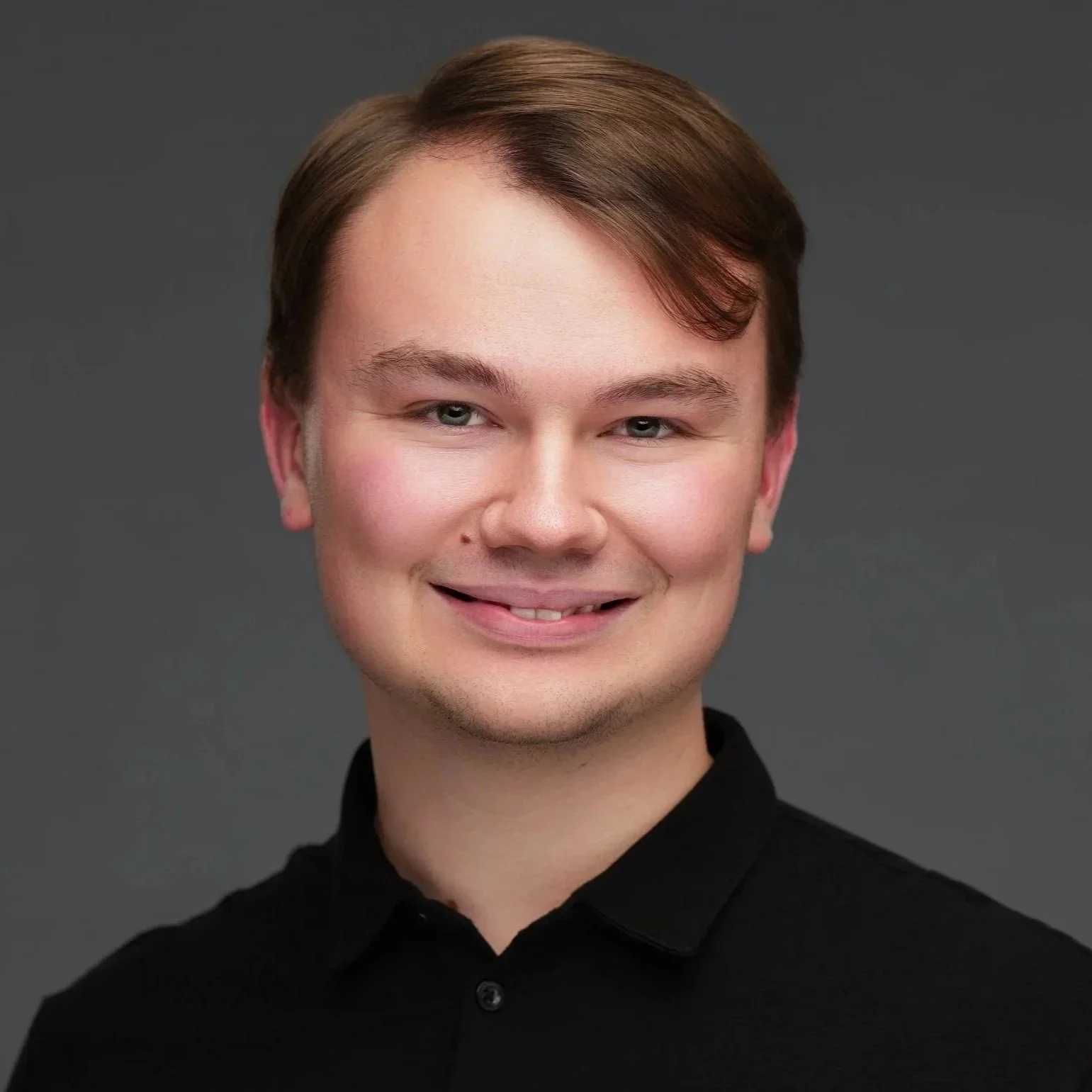 Headshot of a young man with light skin, brown hair, and blue eyes, smiling slightly, wearing a black collared shirt against a gray background.