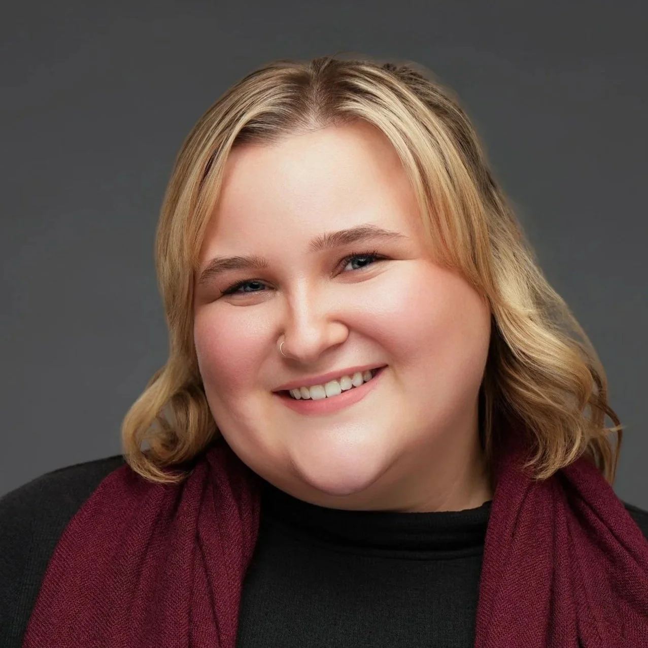 Close-up portrait of a smiling woman with blonde hair, blue eyes, and a nose piercing, wearing a black top and a burgundy vest, against a dark gray background.