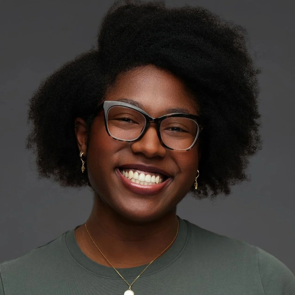 Close-up of a smiling woman with short curly hair, wearing glasses, earrings, a necklace, and a green top against gray background.