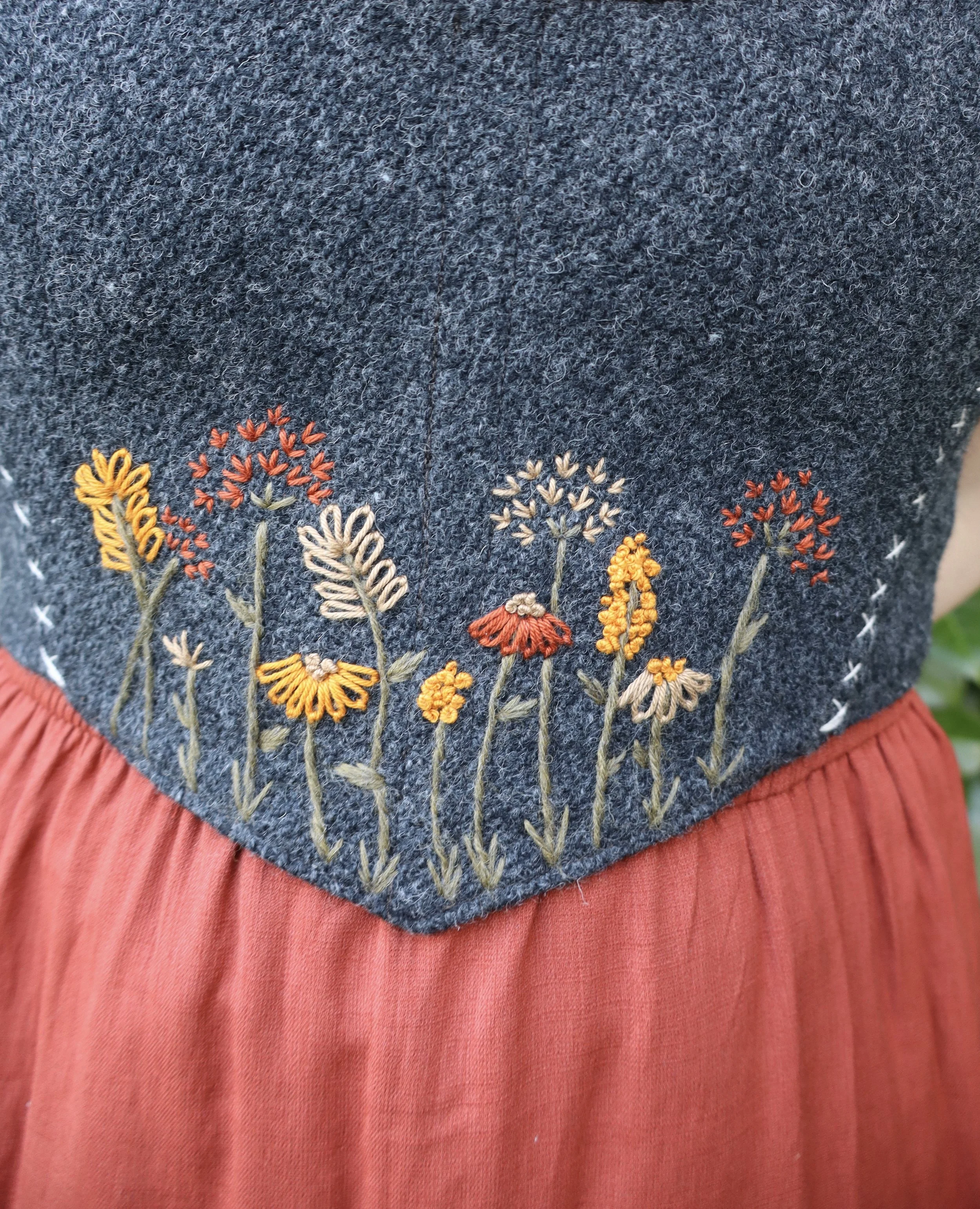 upclose of a woman's midsection wearing a red undershirt and a grey bodice with an embroidered floral motif.