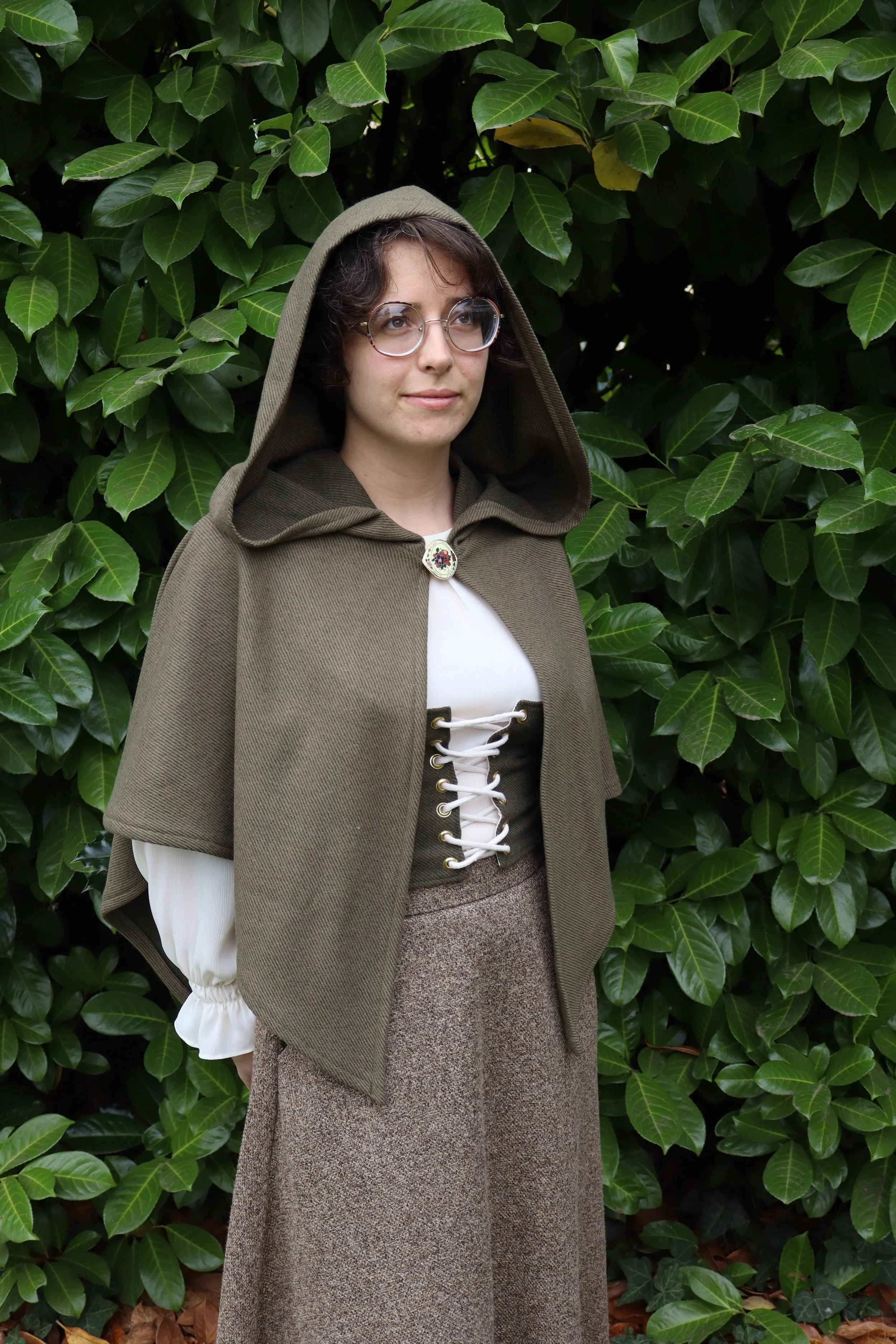 woman with glasses in a dark green capelet/cloak  and a white shirt standing in front of green foliage.