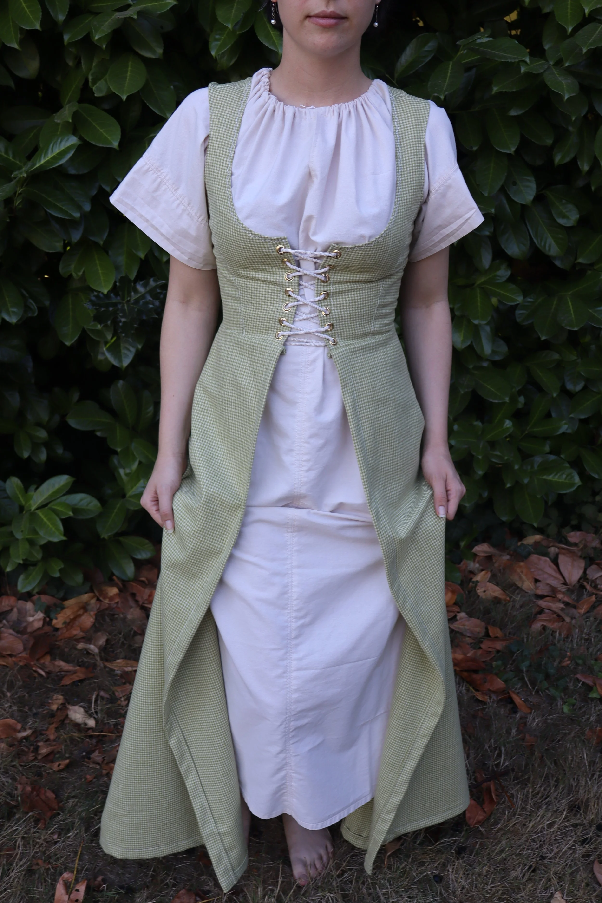 Woman wearing a medieval-style dress and overgown standing outdoors in front of green foliage.