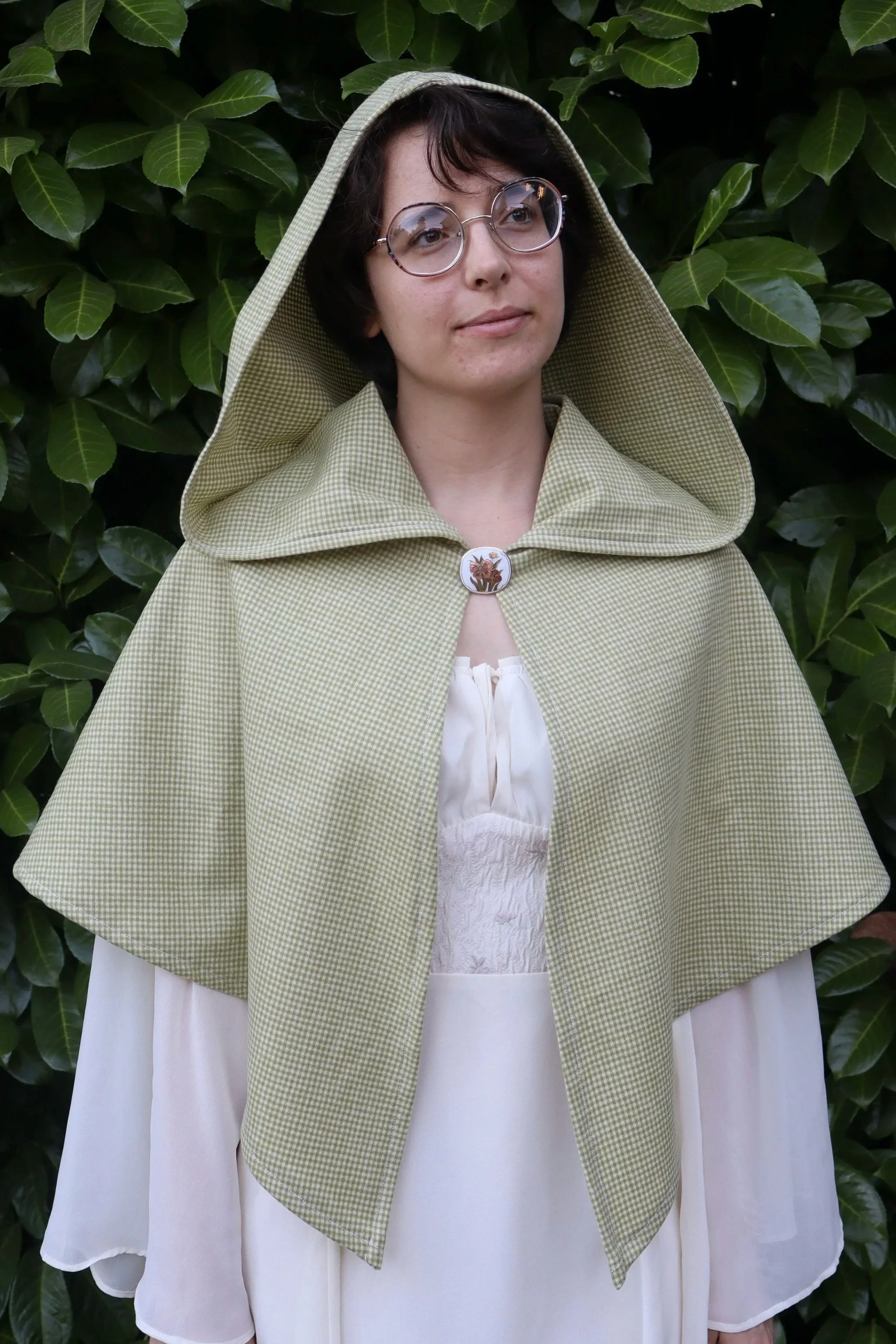 A woman with short dark hair, wearing glasses, a beige hooded cape with a brooch, and a white dress, standing in front of green leafy bushes.