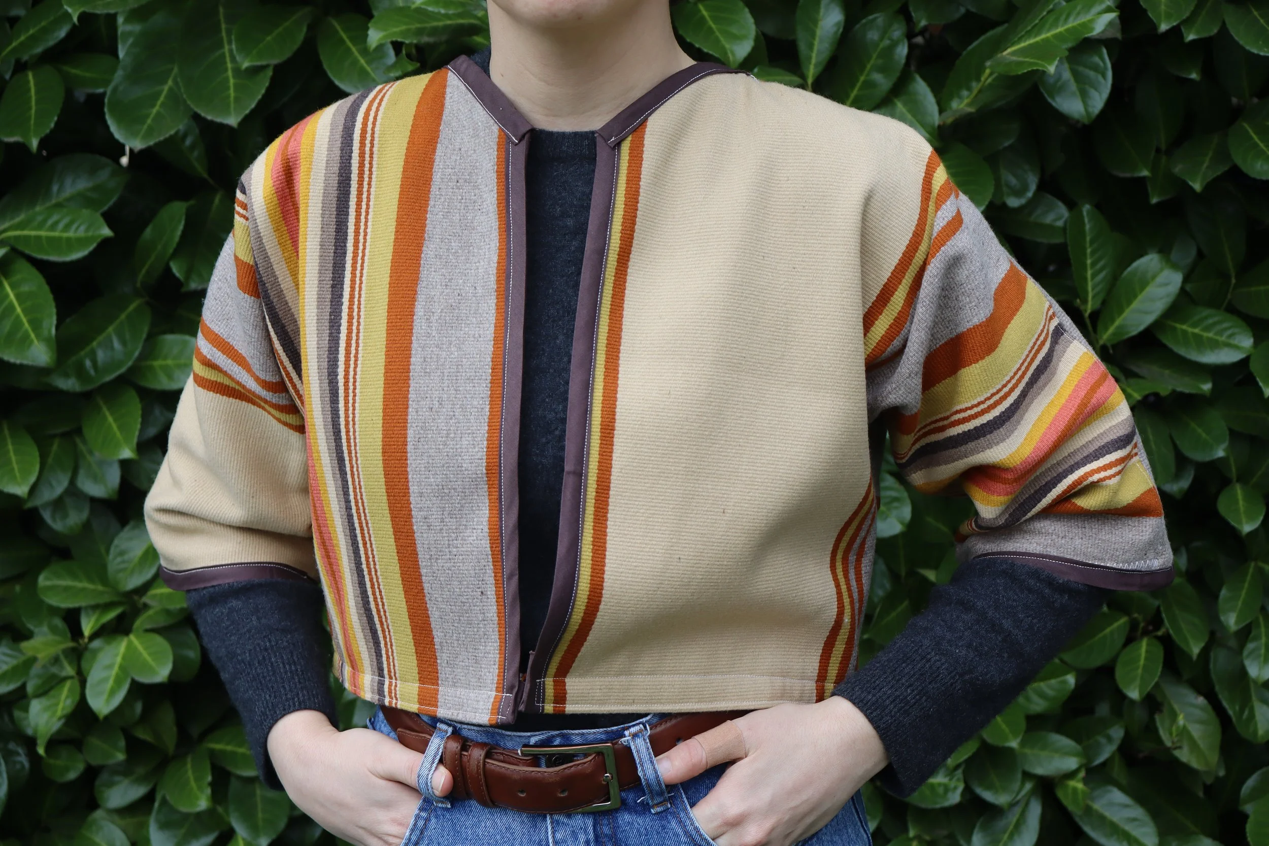 Person wearing a multicolored striped jacket over a dark sweater, standing in front of green leafy background.