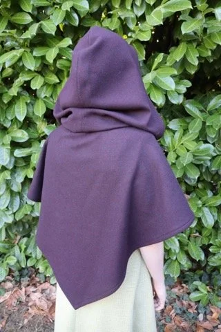 Person wearing a purple hooded cloak standing in front of green leafy bushes.