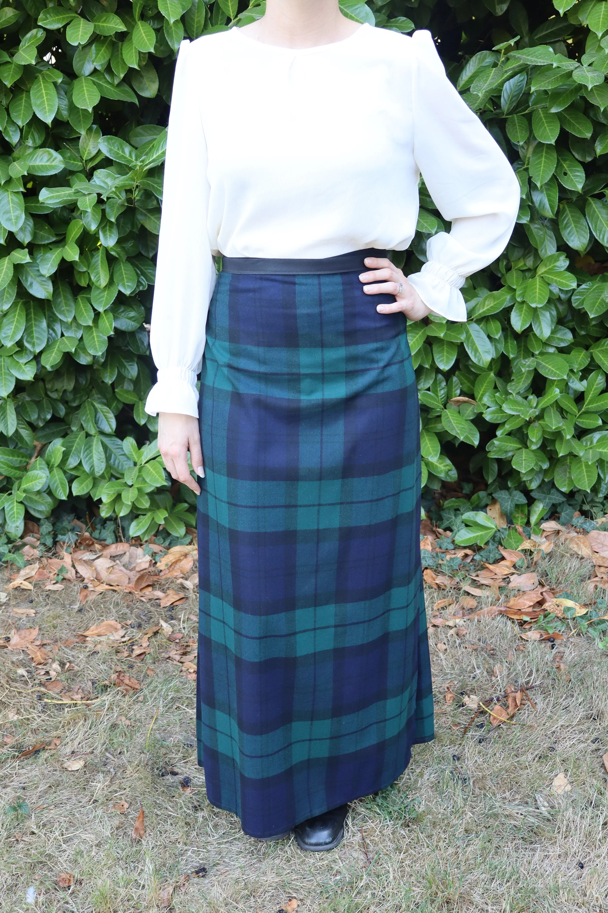 Person wearing a white long-sleeve blouse and a long blue and green plaid skirt, standing outdoors in front of green bushes with fallen leaves on the ground.
