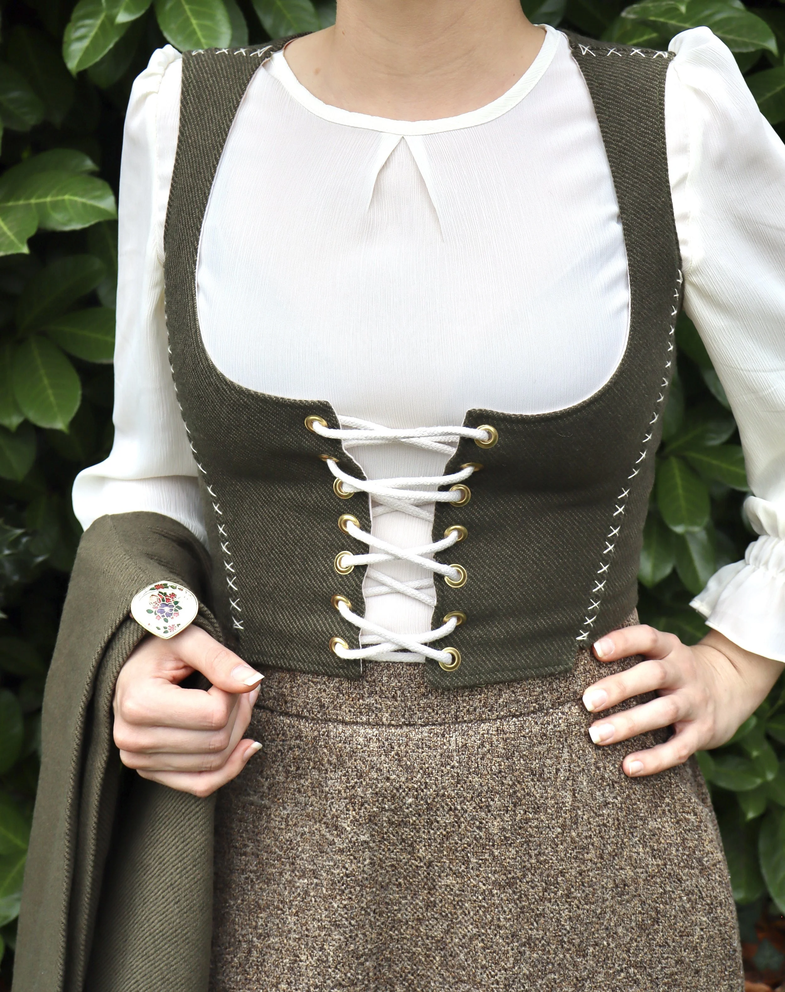 Close-up of a woman wearing a white blouse and a dark green vest with gold eyelets and white laces, holding a green coat or bag, with large green leaves in the background.